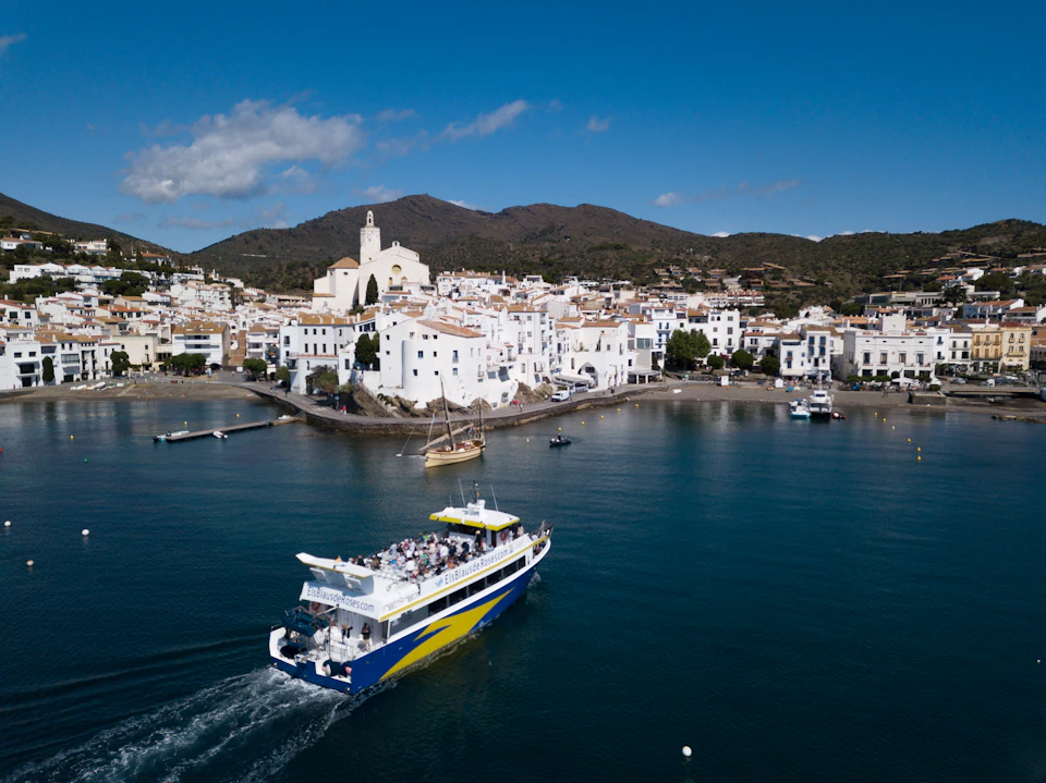 A boat with passengers sails near a coastal town with white buildings, a church, and mountain backdrop.