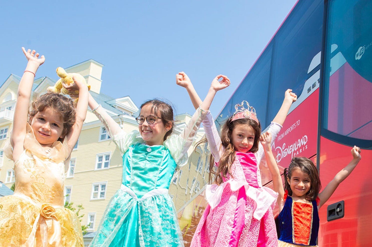 Three children in princess costumes celebrate in front of a red Disney shuttle bus on a sunny day.