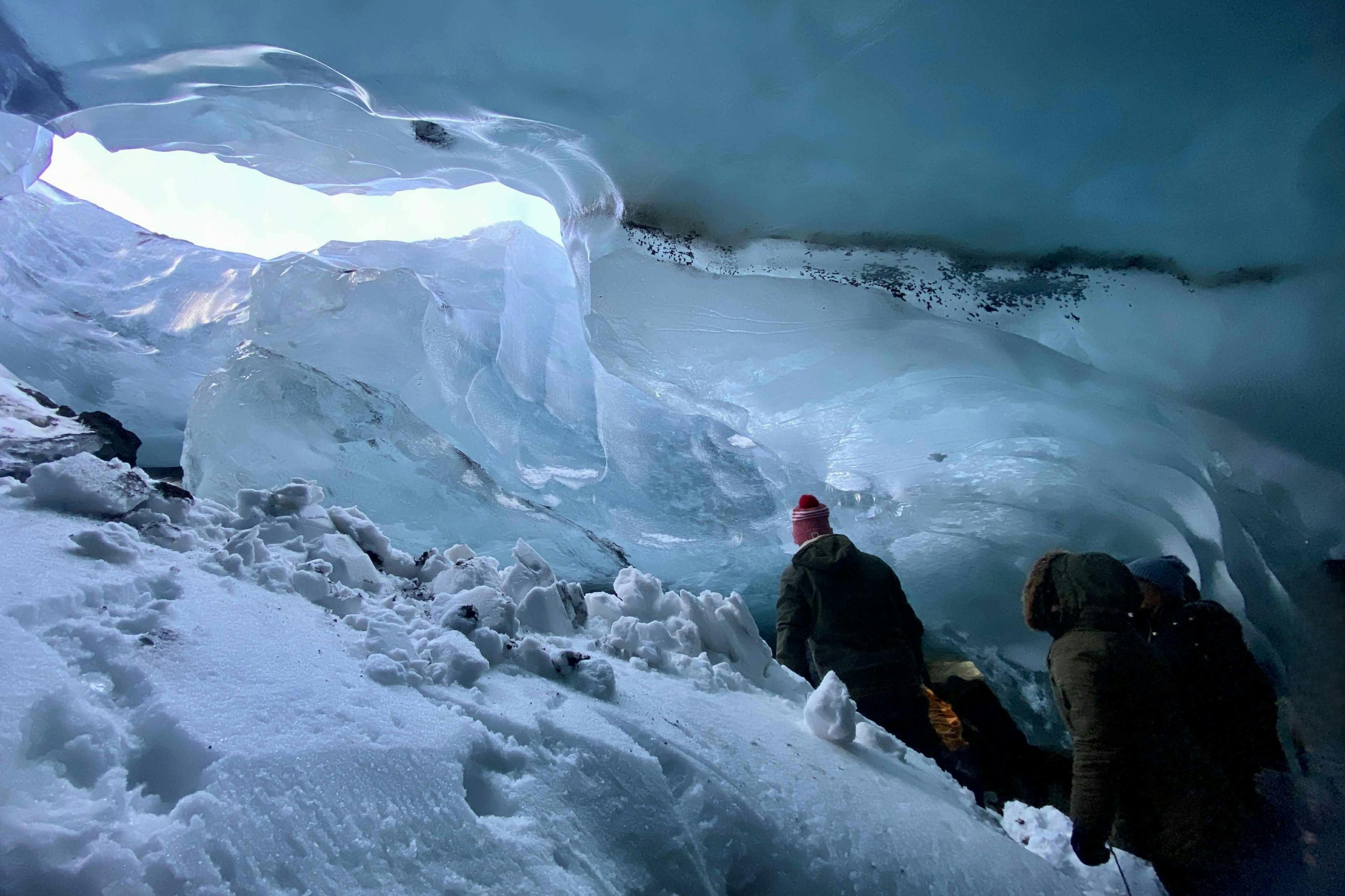 Due persone che indossano abiti invernali esplorano una grotta di ghiaccio blu, con formazioni di neve e ghiaccio che li circondano.