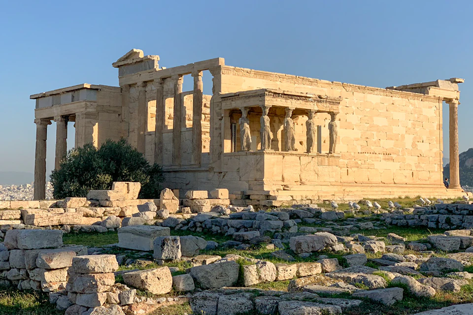 Ancient Greek temple ruins with stone columns and statues, surrounded by scattered rocks and greenery, under a blue sky.