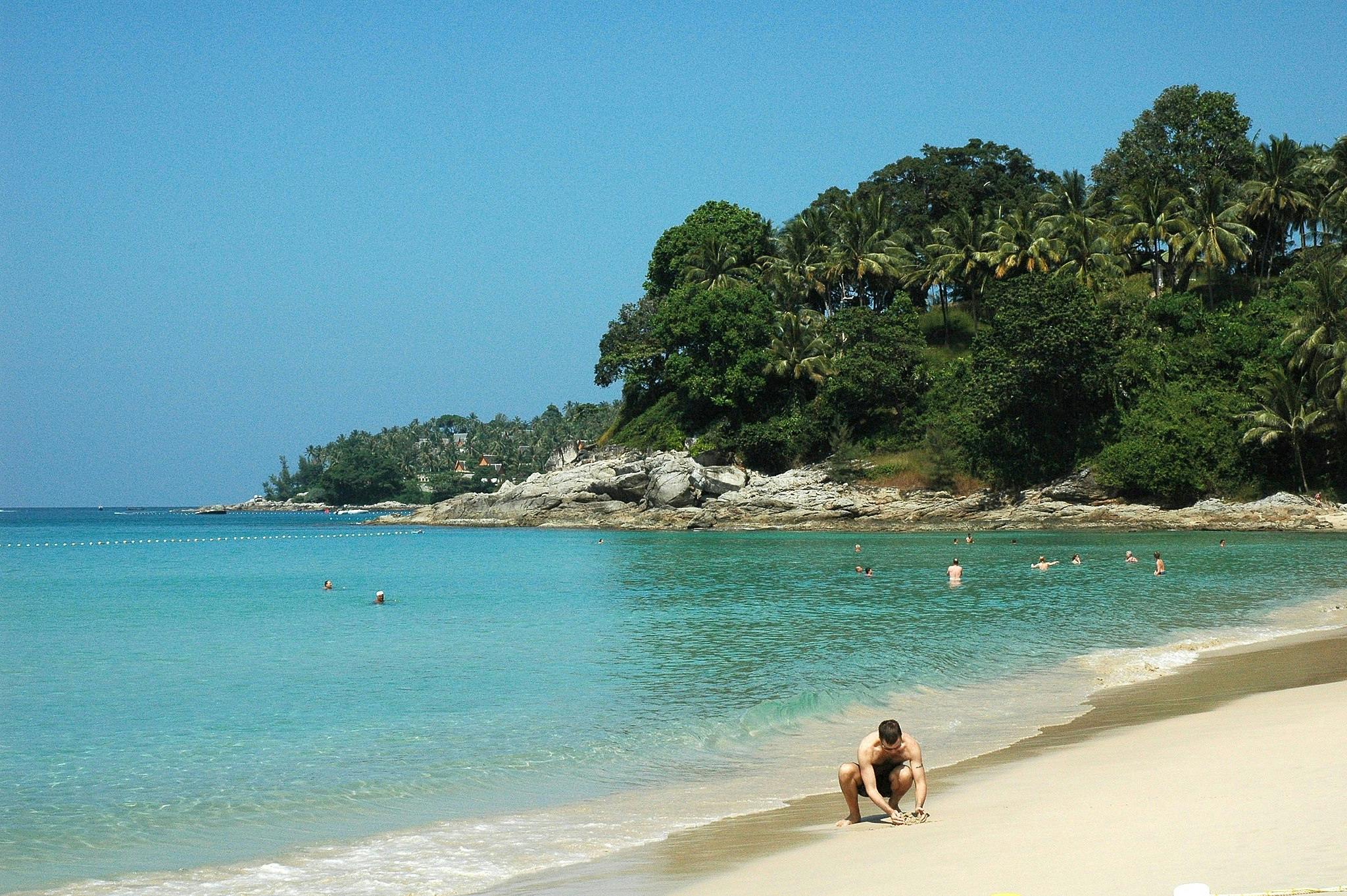 A tropical beach with people swimming in clear blue water. A man in the foreground crouches on the sandy shore. Dense greenery and rocky outcrops in the background.