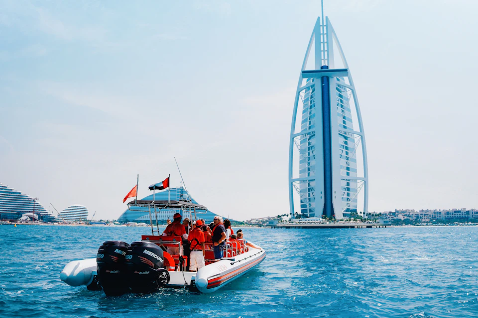 A boat with people in orange life jackets on water near a tall, sail-shaped building and another large boat in the background.