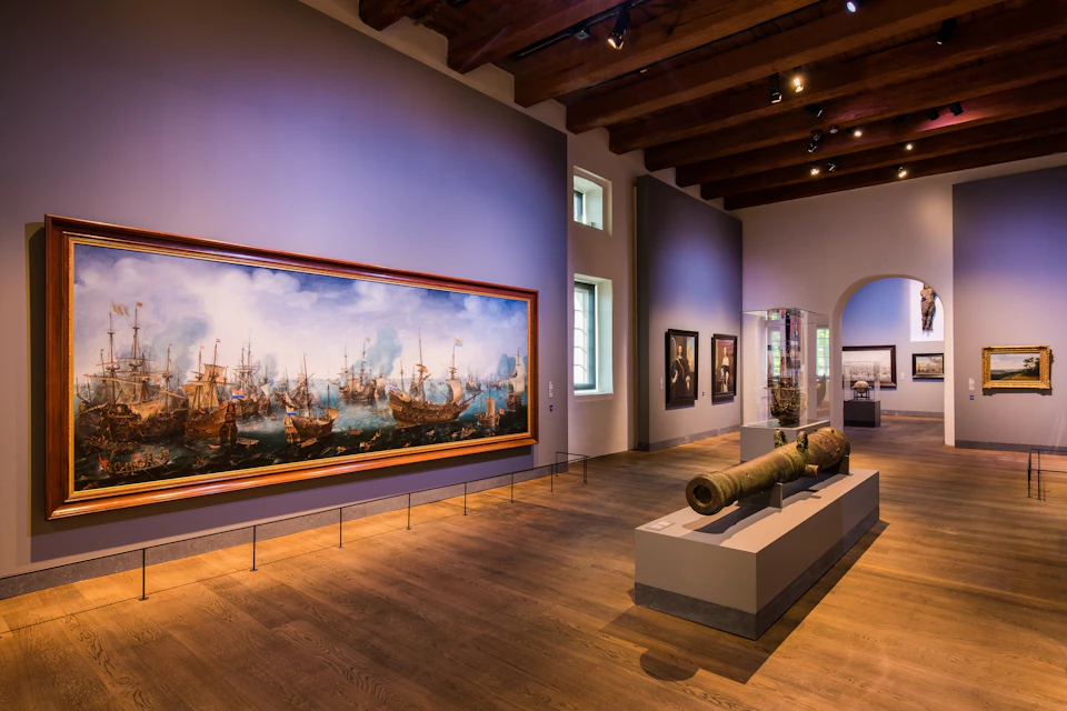 A museum gallery featuring a large seascape painting, antique cannons on display, wooden flooring, and several framed artworks on the walls.