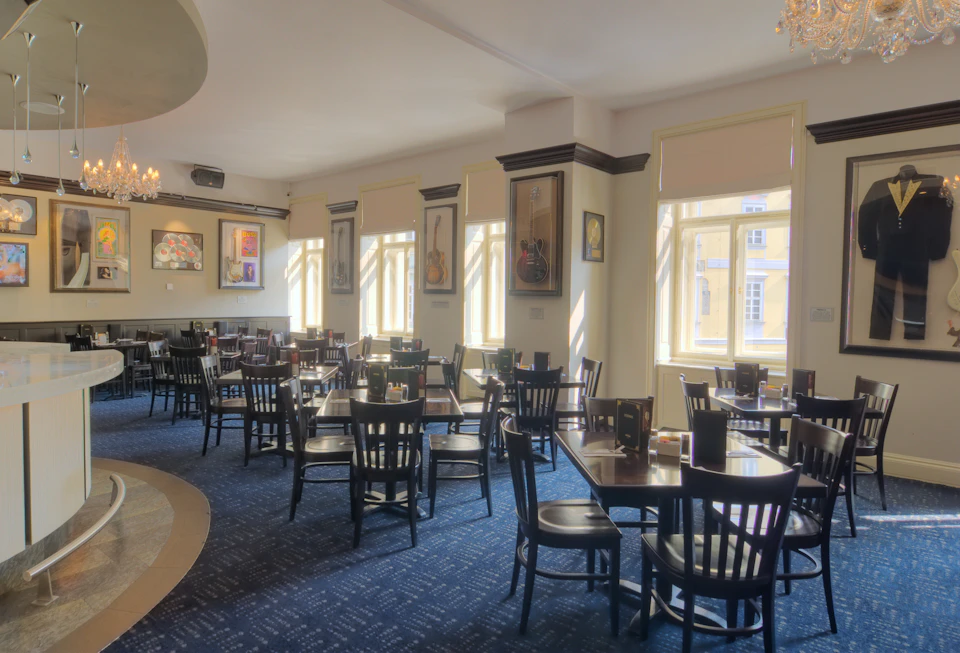 A well-lit, empty restaurant with numerous tables and chairs, framed artworks, and large windows allowing sunlight to stream in.