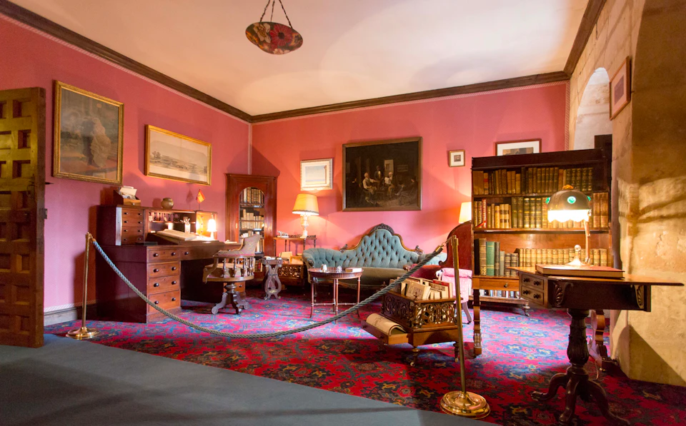 A vintage study room with pink walls, blue sofa, wooden desk, bookshelves, framed paintings, and ornate rug, roped off to visitors.