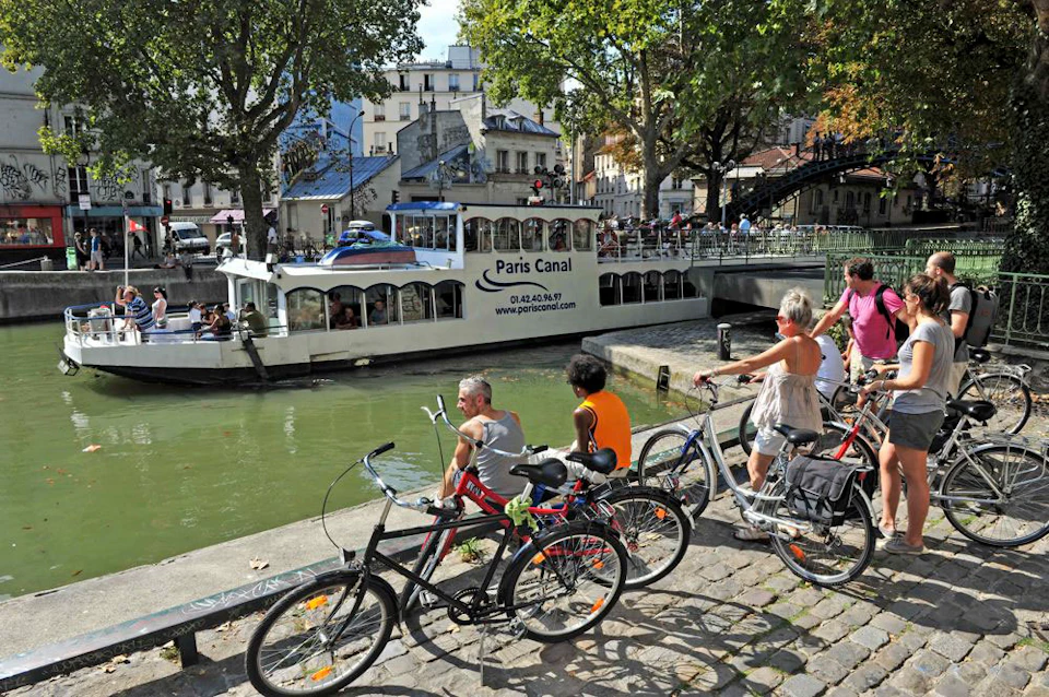 People with bicycles rest by a canal as a tour boat named "Paris Canal" passes by under a bridge; buildings and trees in the background.