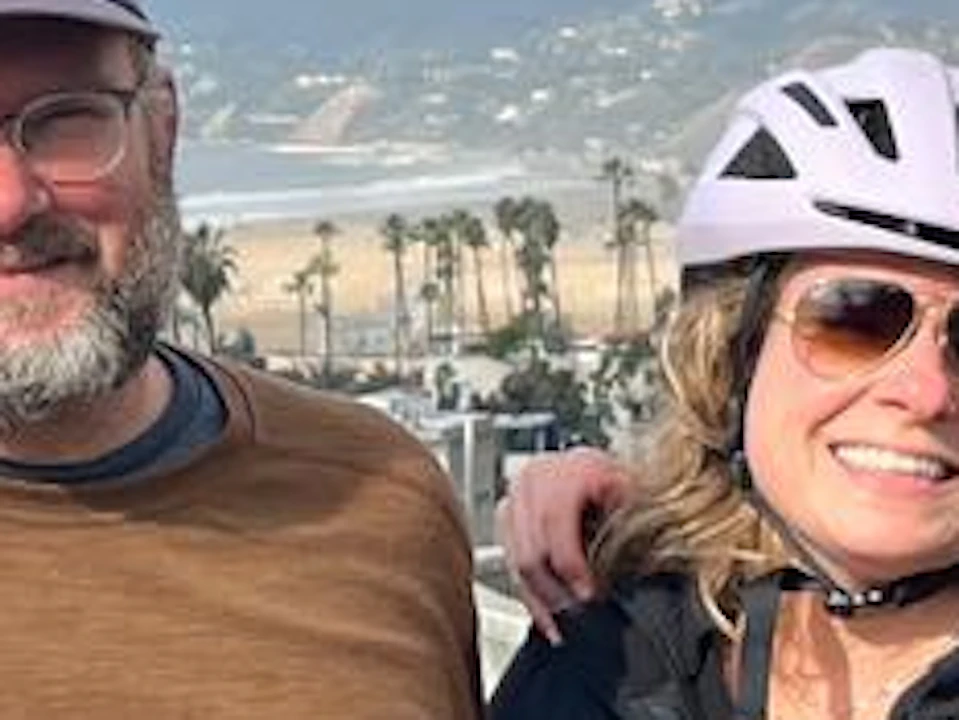 Five people posing by a beach, three with bike helmets. Partly cloudy sky, ocean, and palm trees in the background.