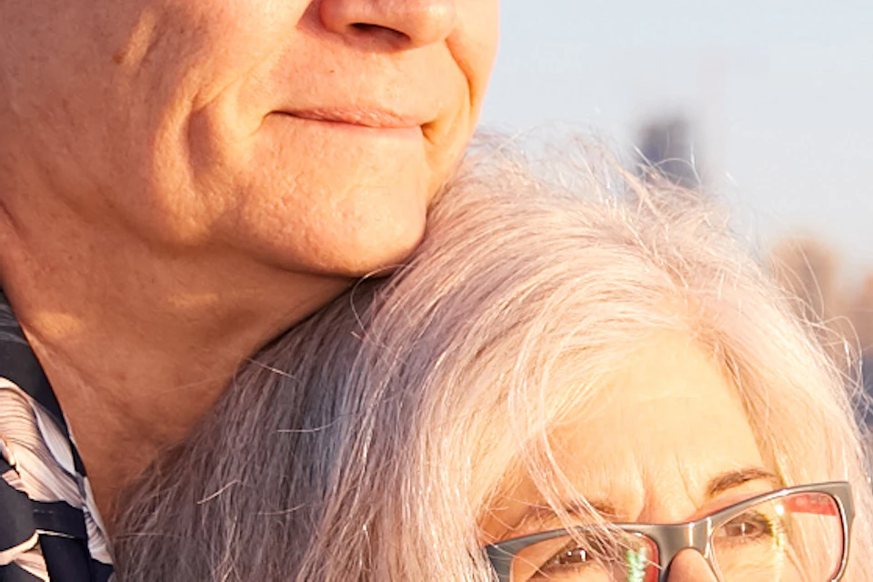 An elderly couple enjoys a boat ride, standing close together with a city skyline in the background. The man wears sunglasses.