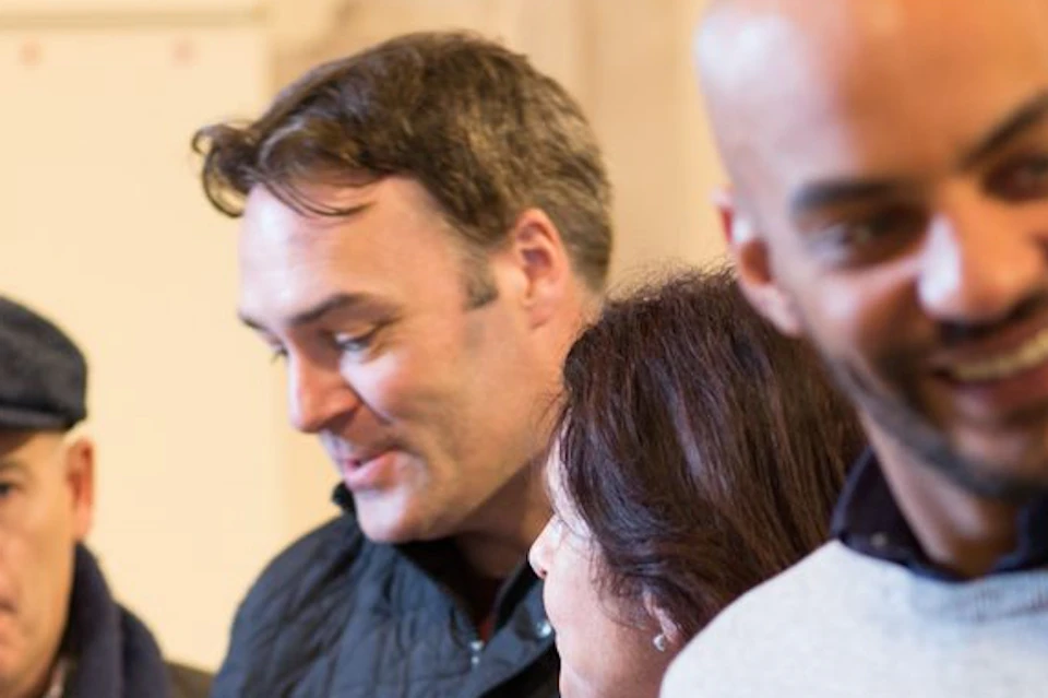 People standing at a counter, holding glasses of whiskey, engaged in conversation. One person wears a cap; another smiles.