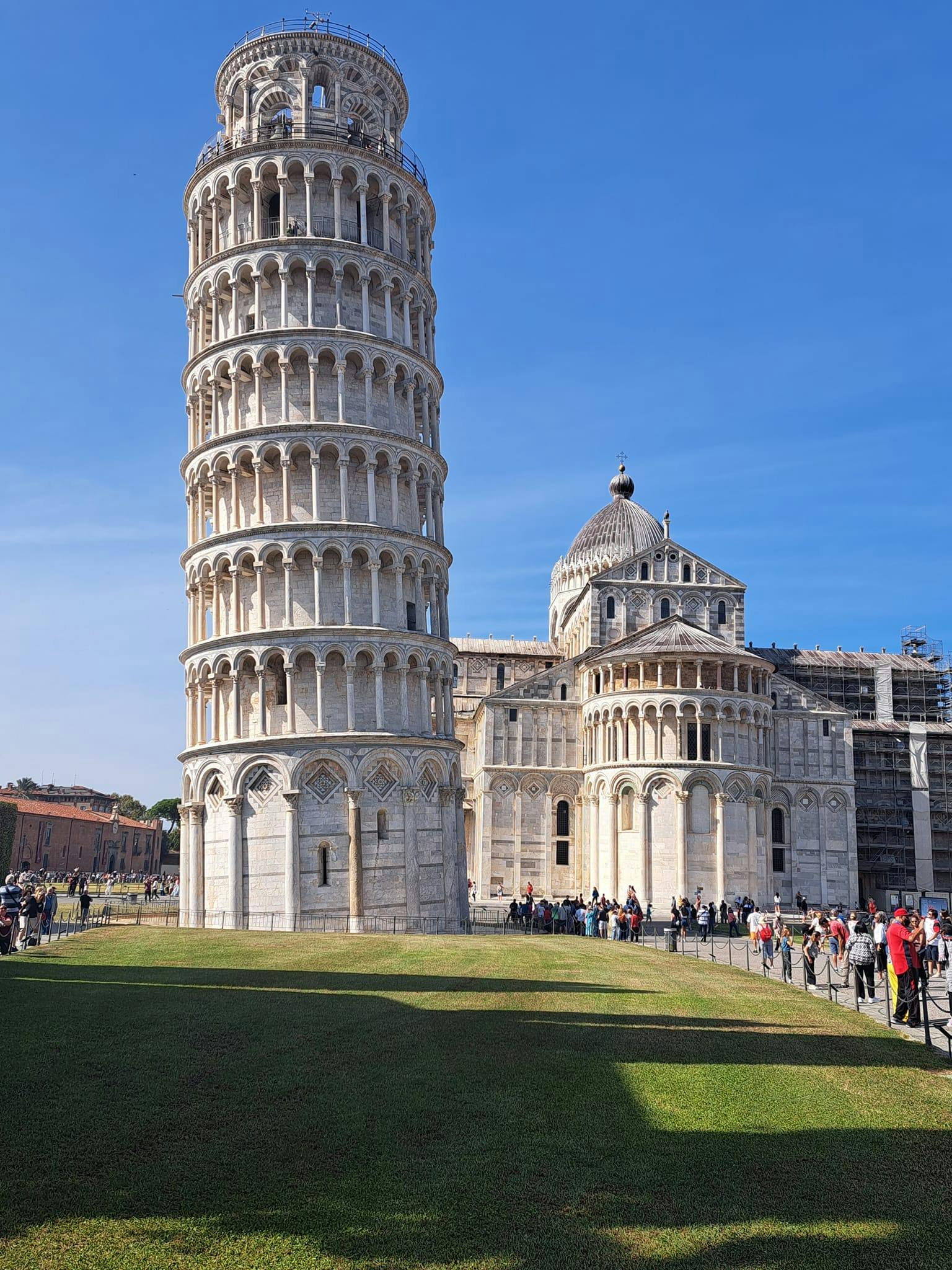 Ο κεκλιμένος πύργος της Πίζας και ο καθεδρικός ναός της Πίζας κάτω από έναν καταγάλανο ουρανό, με τους ανθρώπους να συγκεντρώνονται γύρω από το γρασίδι και το διάδρομο.