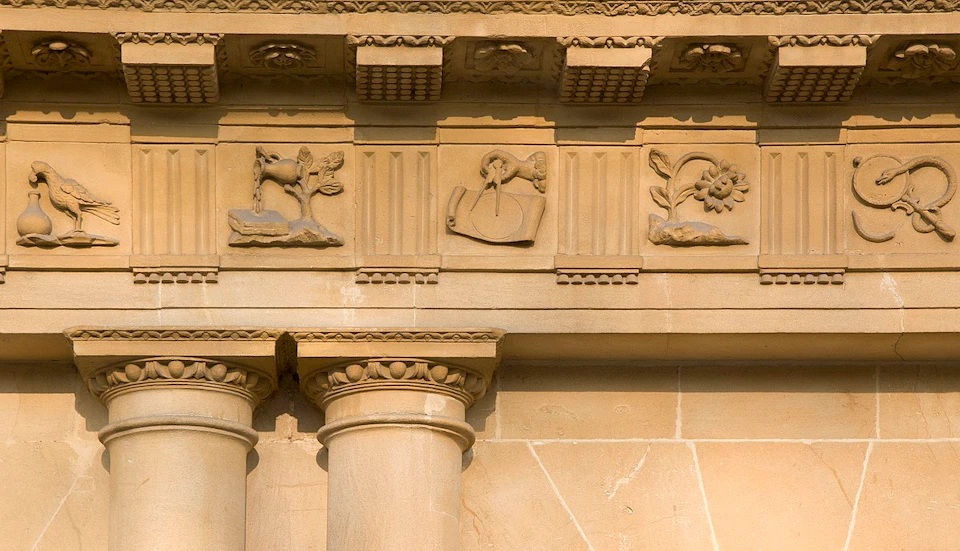 Ornate stone architecture with columns and carvings featuring a goat, a sun dial, and a flower.
