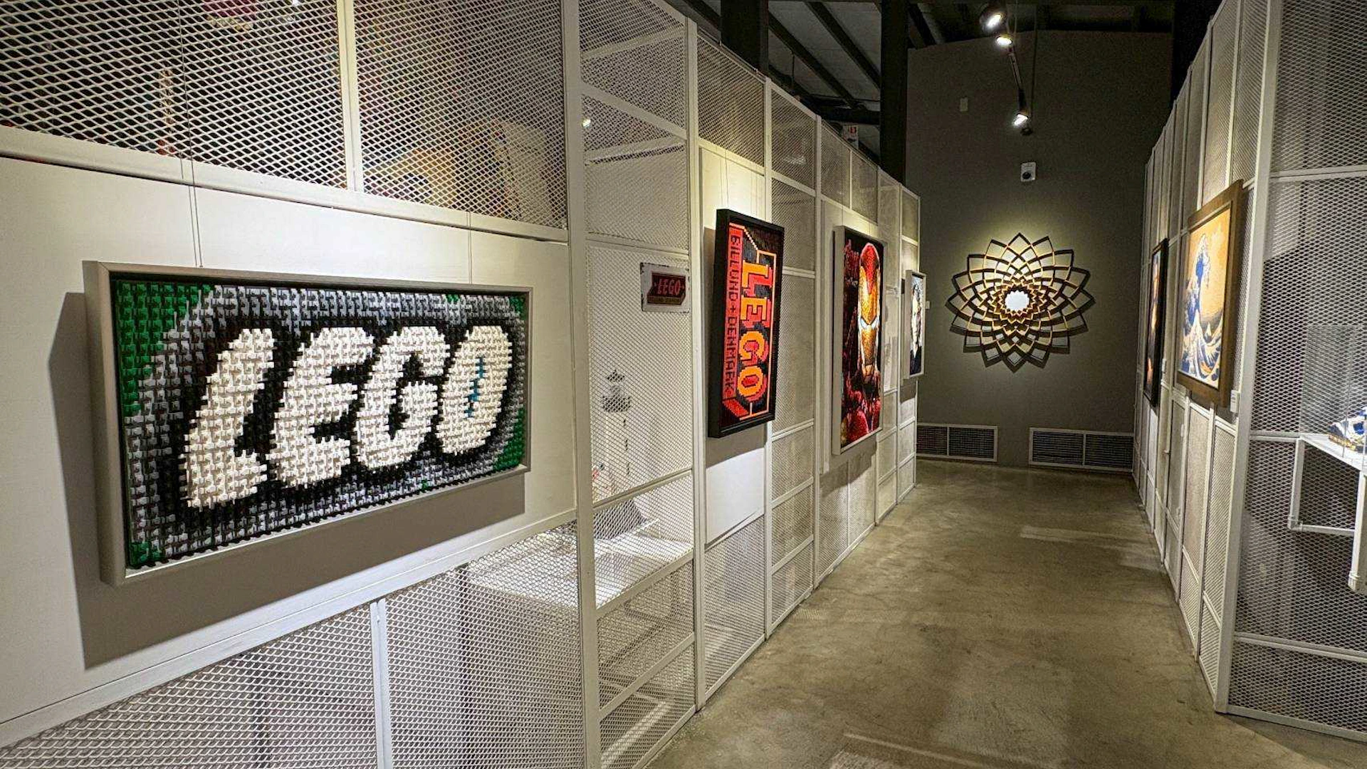 A gallery display featuring framed LEGO artwork on a perforated white wall with decorative lighting and a geometric wall piece.