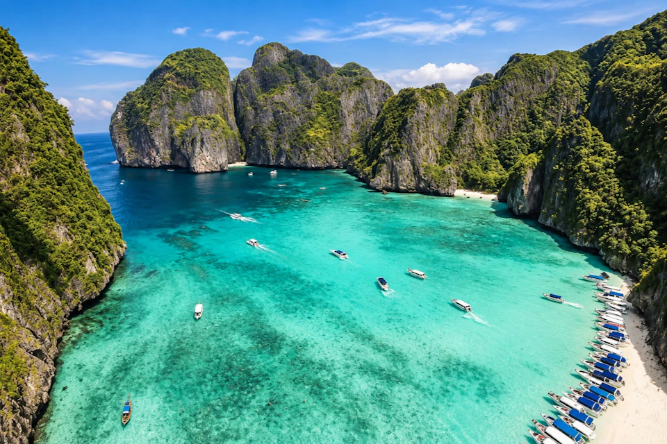 A tropical bay with turquoise water, boats scattered across, and surrounded by lush, rocky cliffs under a blue sky.