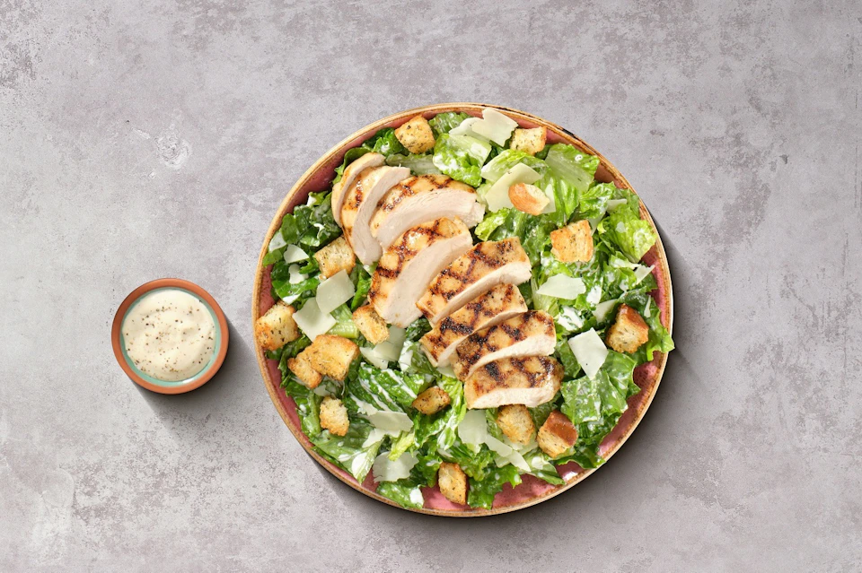 A Caesar salad with grilled chicken slices, croutons, and Parmesan cheese served in a bowl, with a side of dressing in a small dish.