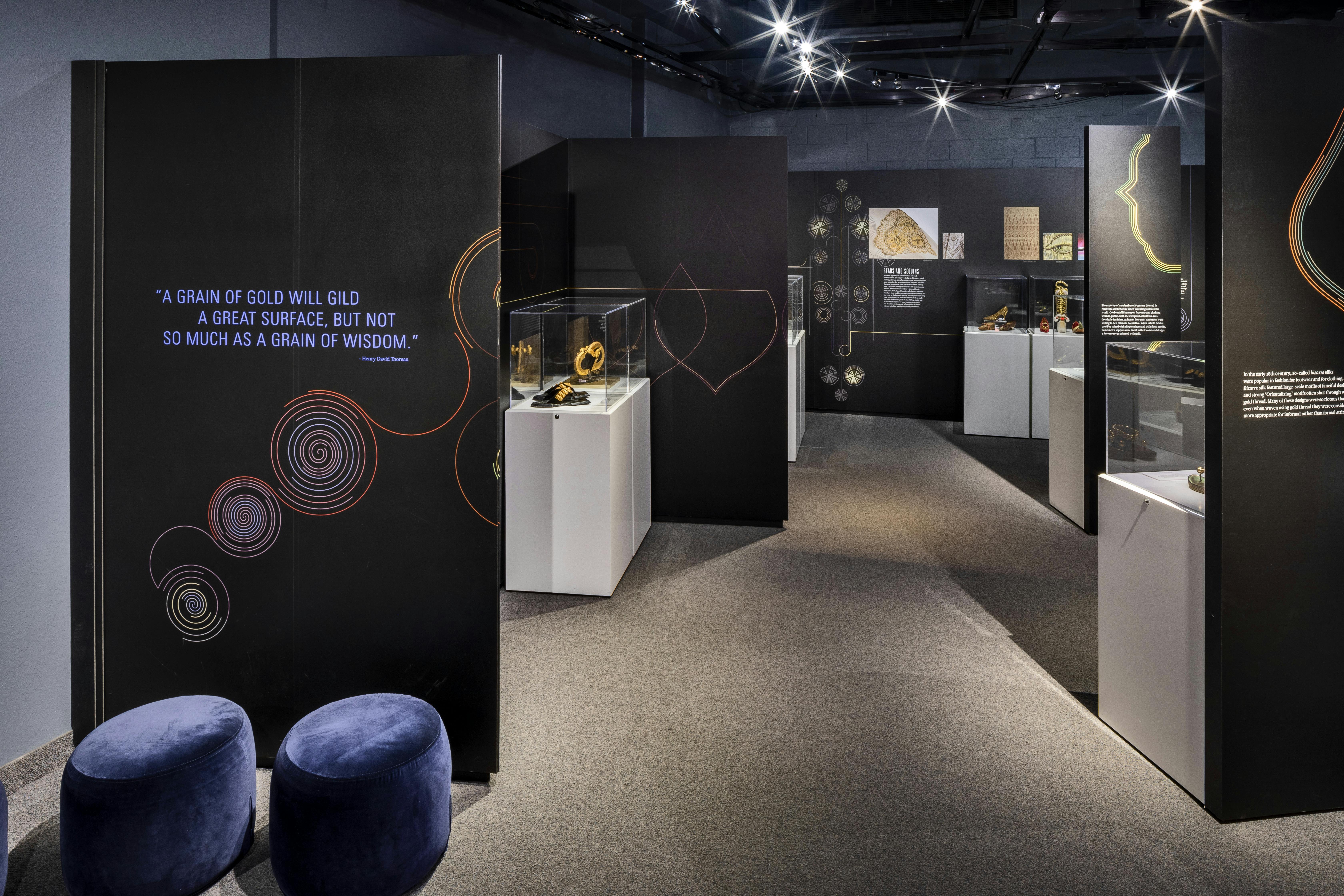 Moderne museumsudstilling med artefakter i glaskasser, sorte vægge med guld og rødt design og blå fløjlsstole.