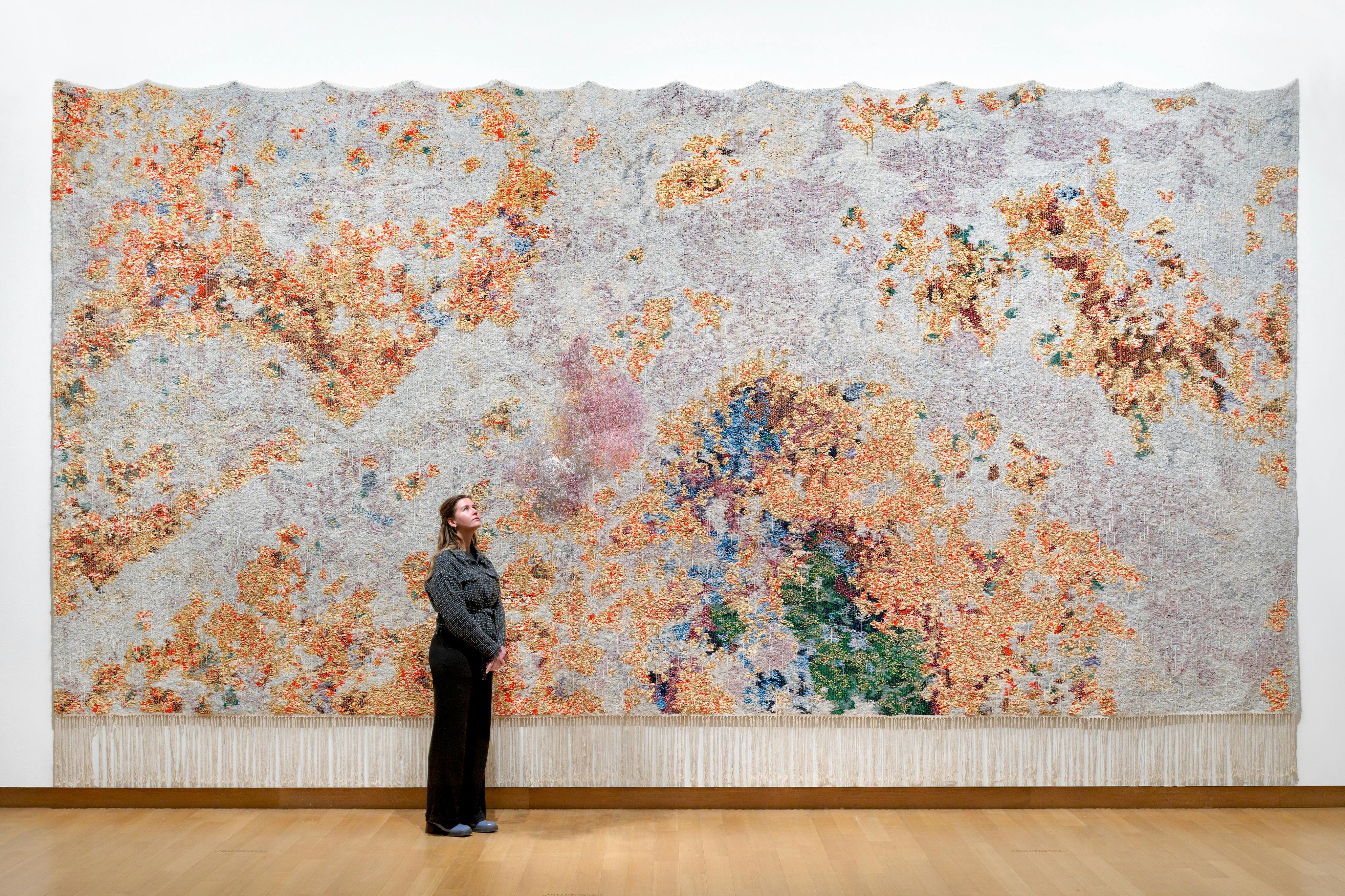 Uma pessoa está em frente a uma obra de arte têxtil grande e colorida exposta numa parede de uma galeria.