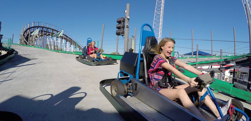 A young girl drives a go-kart on a race track, smiling with excitement under a clear blue sky.