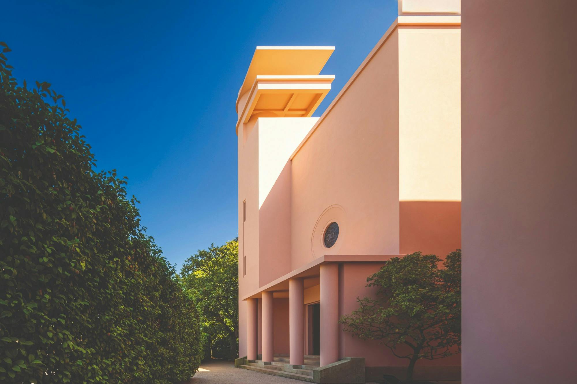 A pastel pink building with columns and a unique architectural design stands beside a hedge under a clear blue sky.