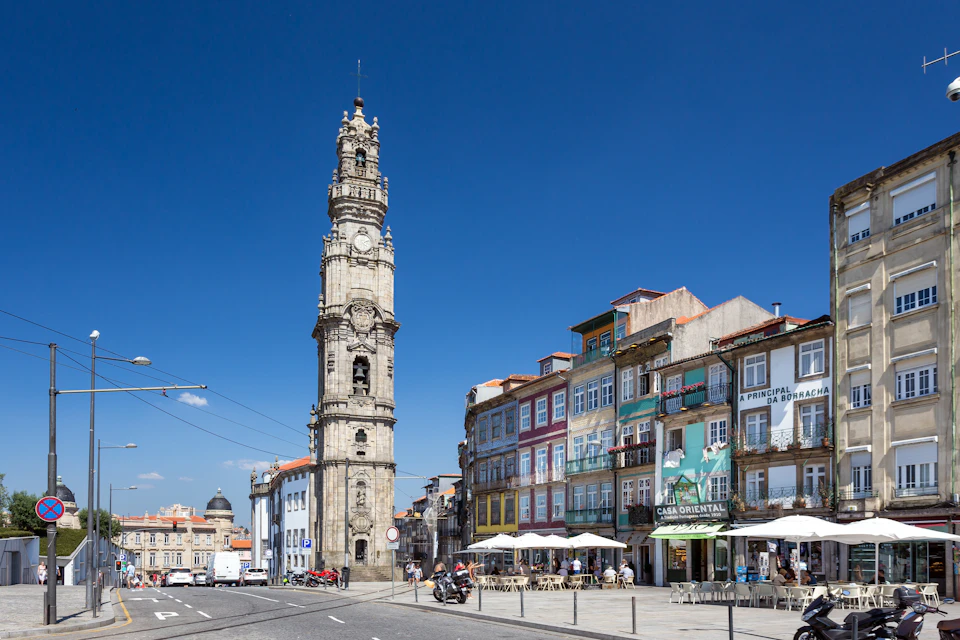 Stone bell tower beside colorful multi-story buildings with outdoor café seating under white umbrellas on a clear, sunny day.
