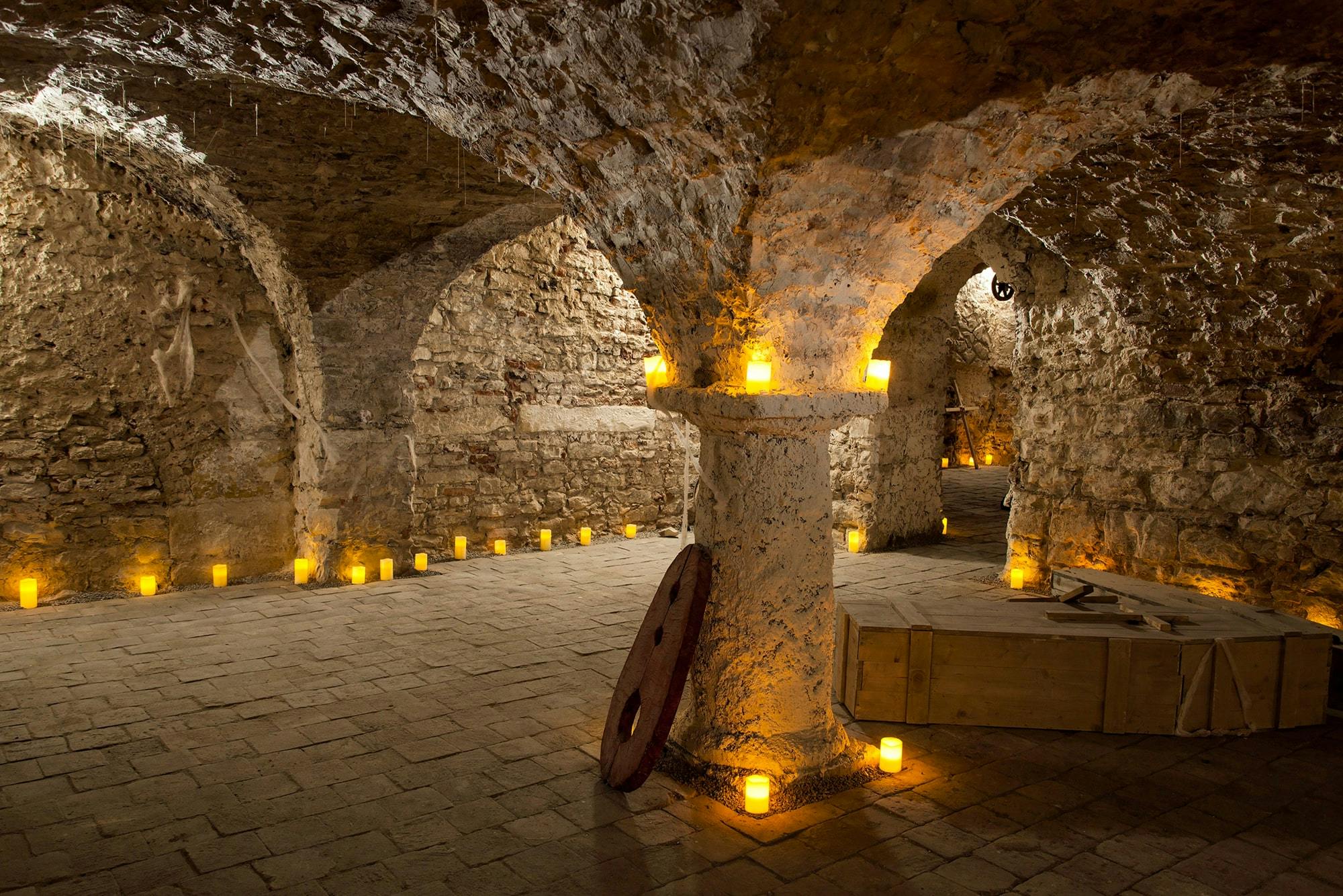 Ένα αμυδρά φωτισμένο πέτρινο κελάρι με τοξωτά ταβάνια, κεριά που καλύπτουν το πάτωμα, μια ξύλινη ρόδα καροτσιού και ένα ξύλινο κιβώτιο.