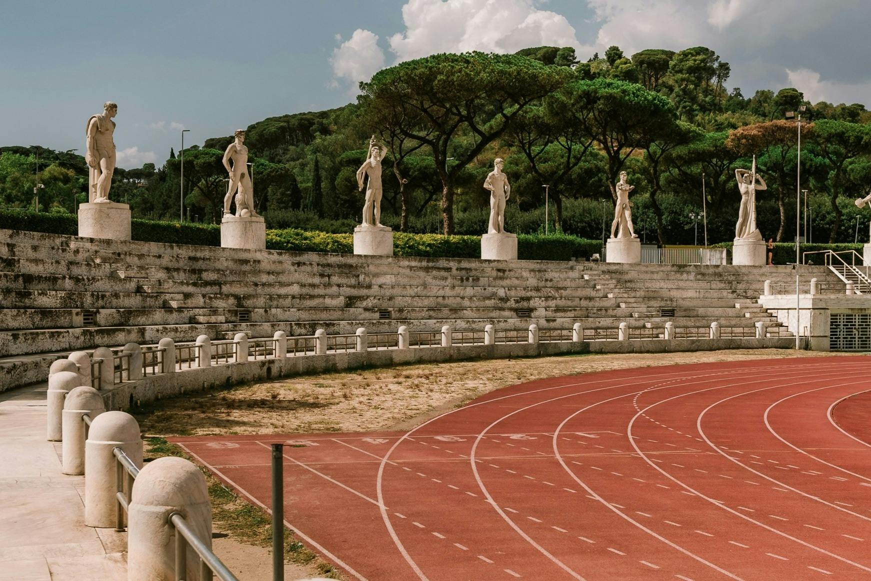 Een atletiekbaan maakt bochten voor stenen stadionstoelen met vier standbeelden en bomen op de achtergrond onder een gedeeltelijk bewolkte hemel.
