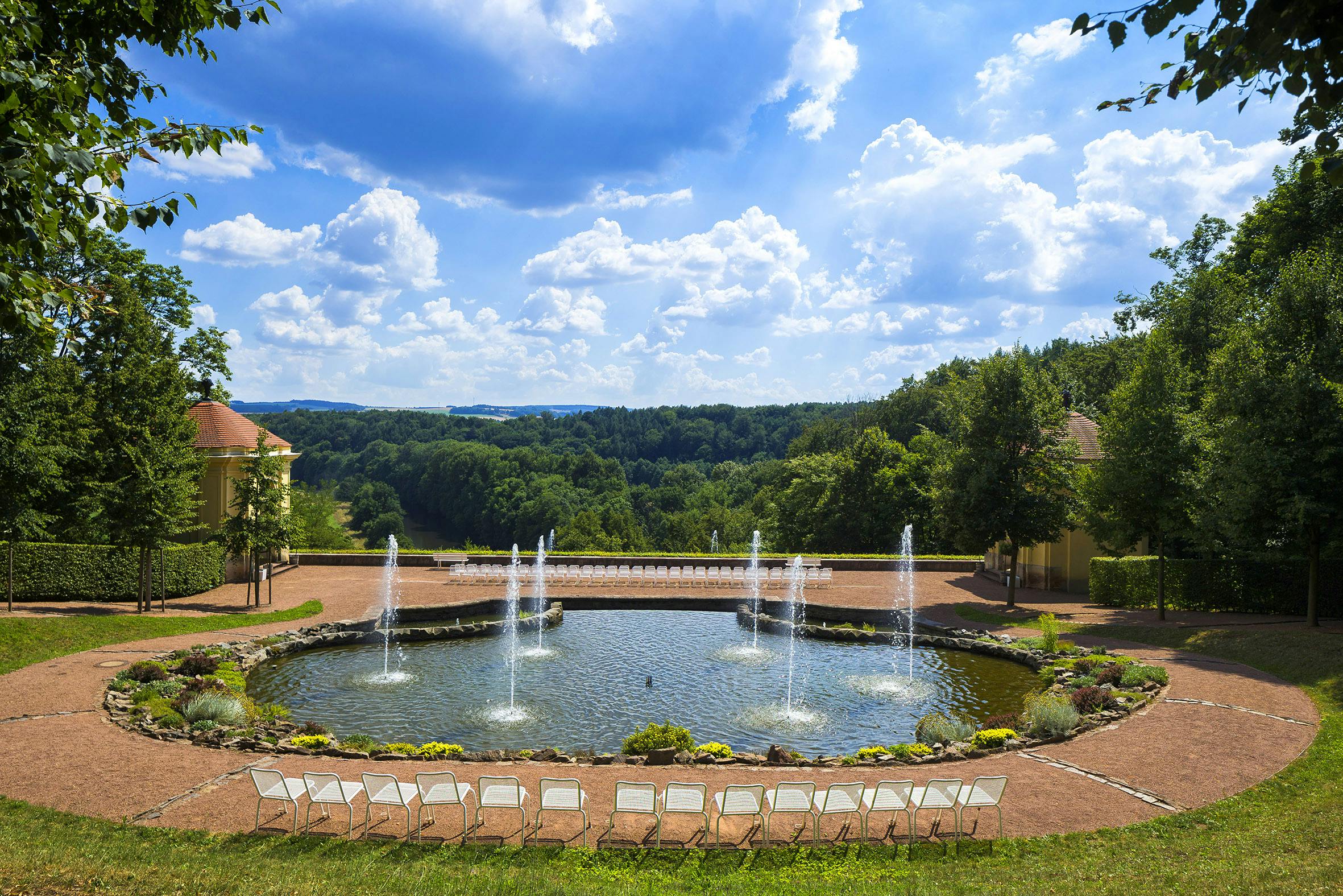 Una veduta panoramica presenta una fontana in uno stagno, una fila di sedie vuote in primo piano e un paesaggio boscoso sotto un cielo azzurro con nuvole.