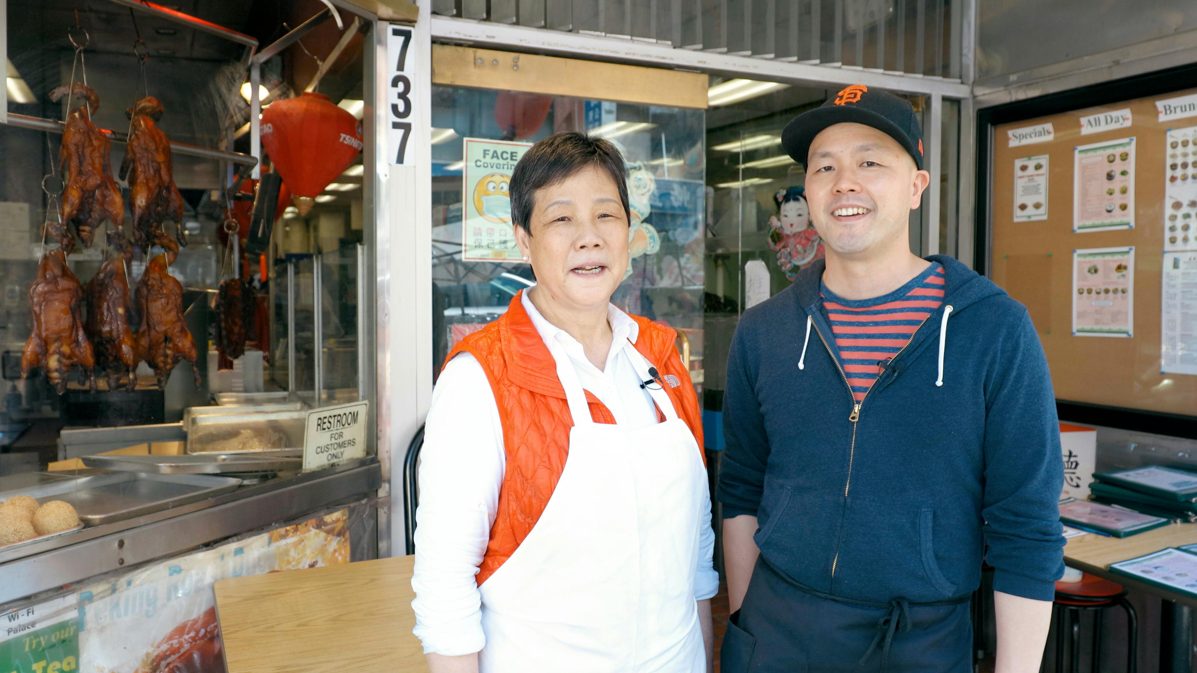 Twee mensen glimlachen voor een winkelpui. De een draagt een schort en rood vest, de ander een capuchon en zwarte pet.