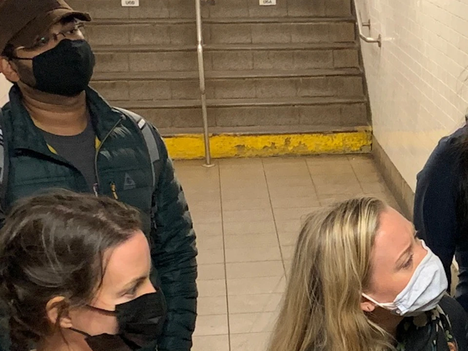 A group of people wearing masks stand in a subway station, examining a wall panel with buttons and a screen.
