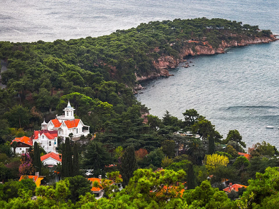 A coastal scene with a large white and red-roofed building surrounded by trees, and a rocky shoreline leading to the sea.