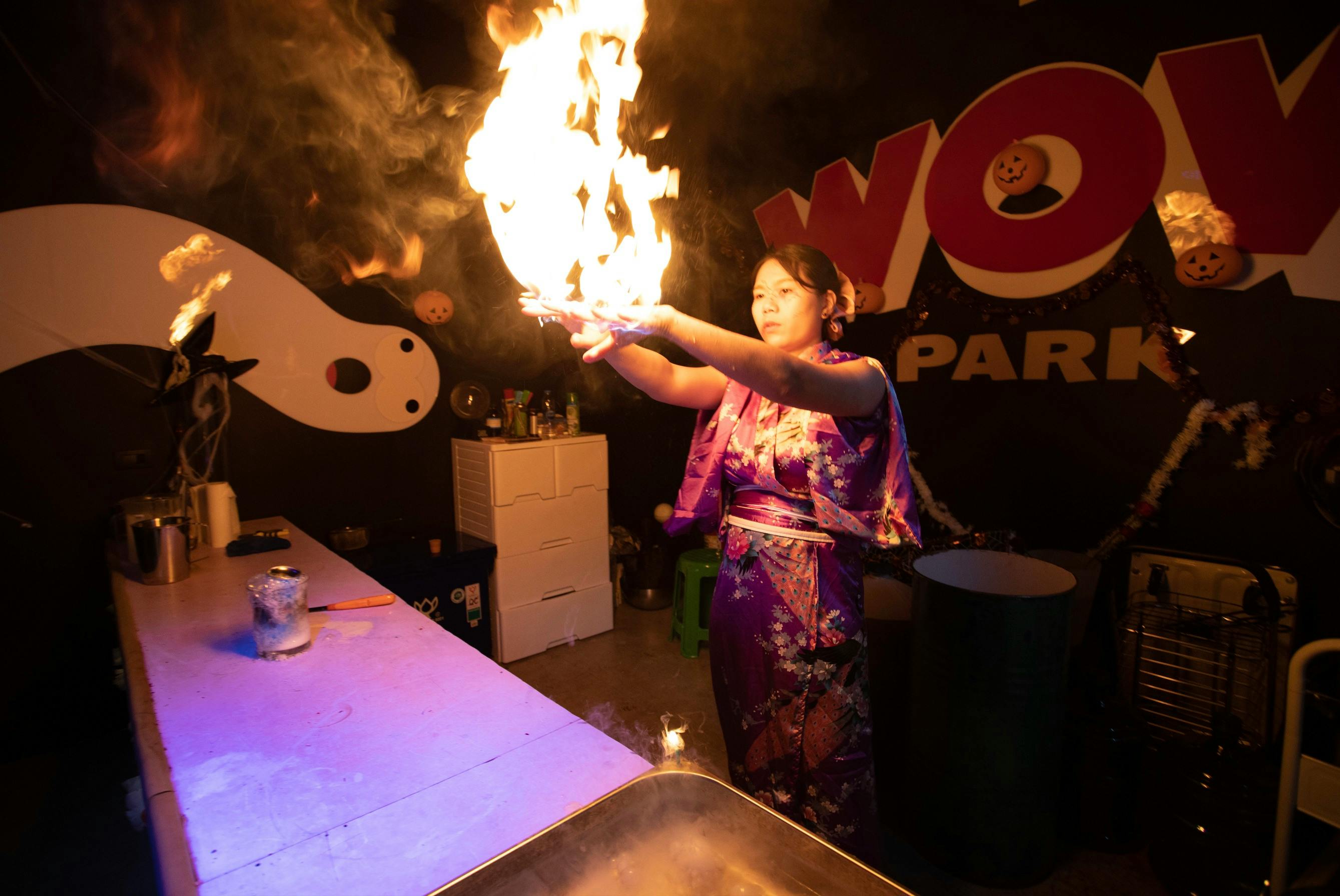 A person wearing a traditional outfit performs a fire act indoors, with a flame rising from their hand.