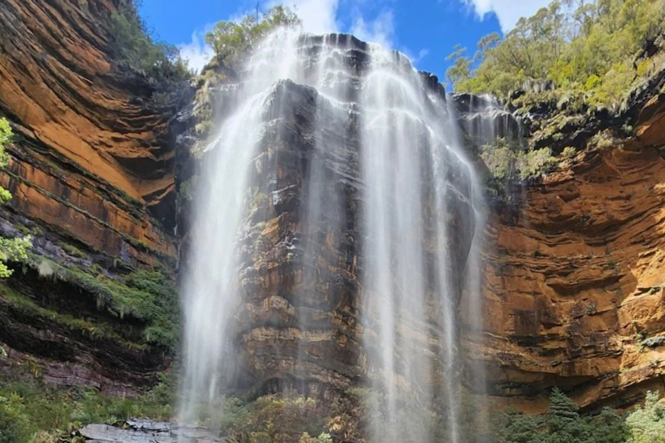 A tall waterfall cascades down a rugged cliff surrounded by lush vegetation under a bright blue sky.