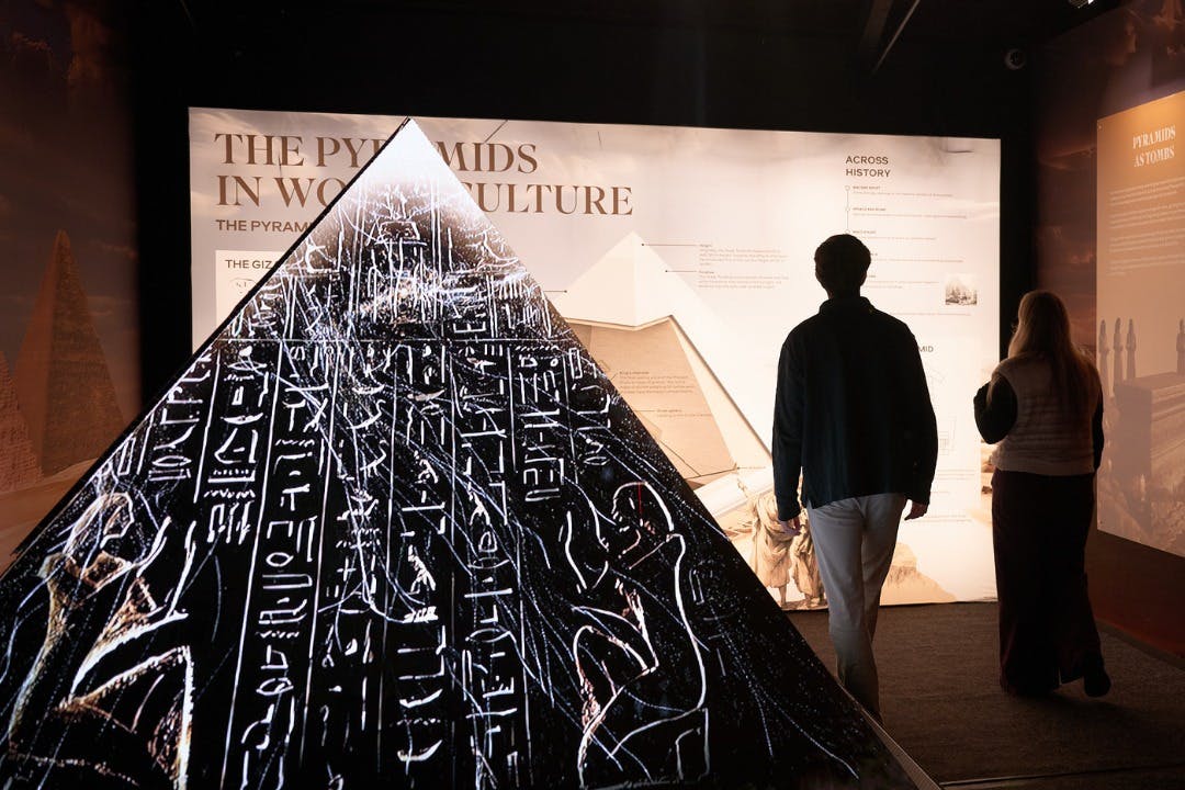 Utställning med en pyramidmodell med hieroglyfer och besökare som observerar informationspaneler i ett svagt upplyst rum.