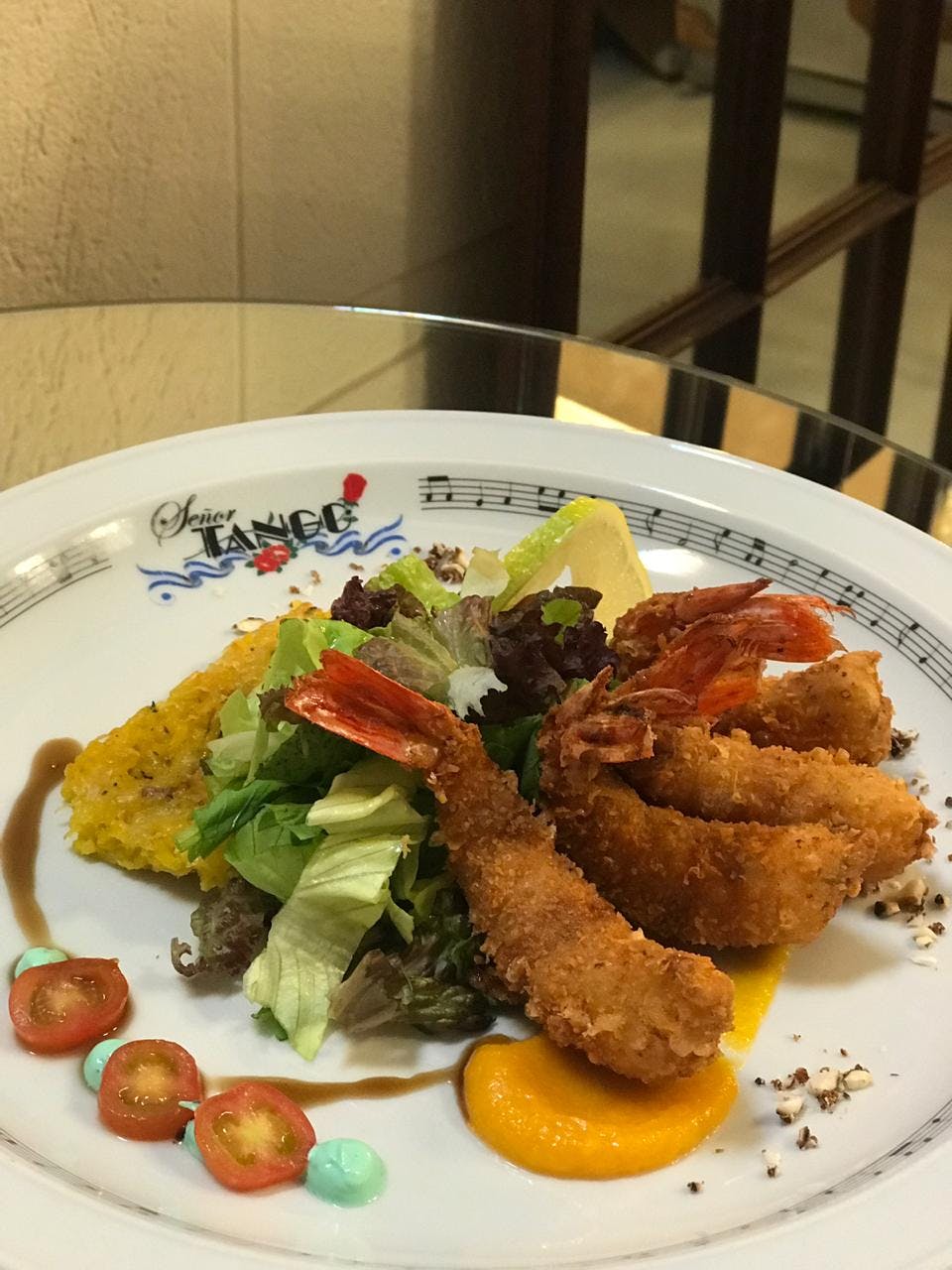 A plate of salad, fried shrimp, squash, and tomato slices, garnished with sauce and served in a decorated plate.