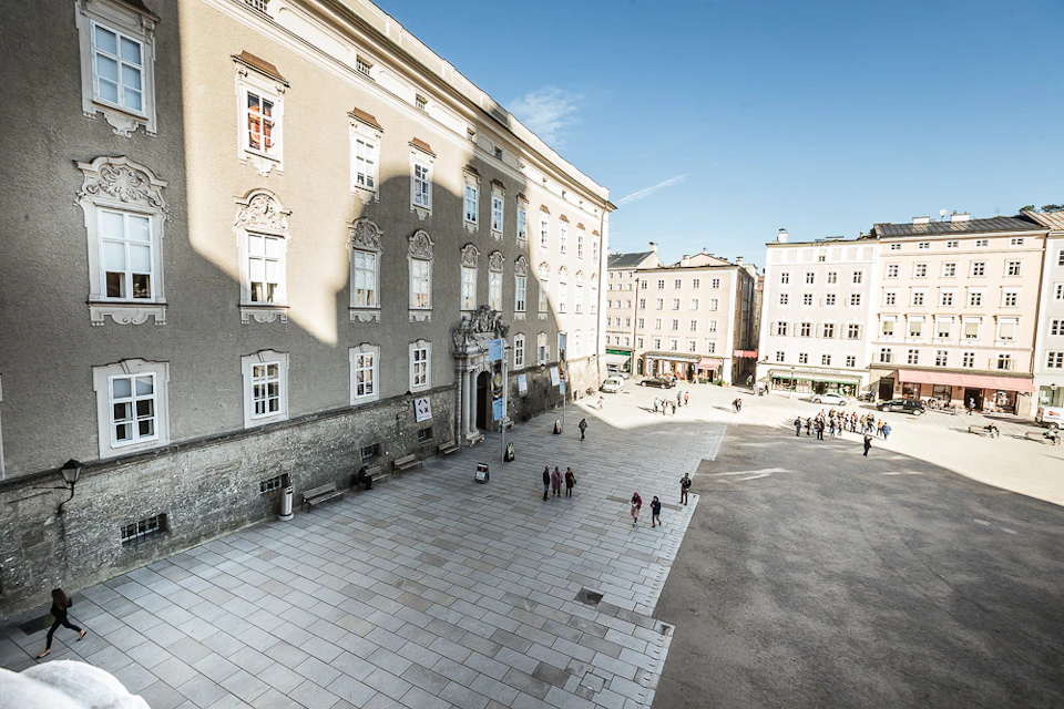 A sunny square in front of a historic building, with people walking about and small groups gathered, under a clear blue sky.