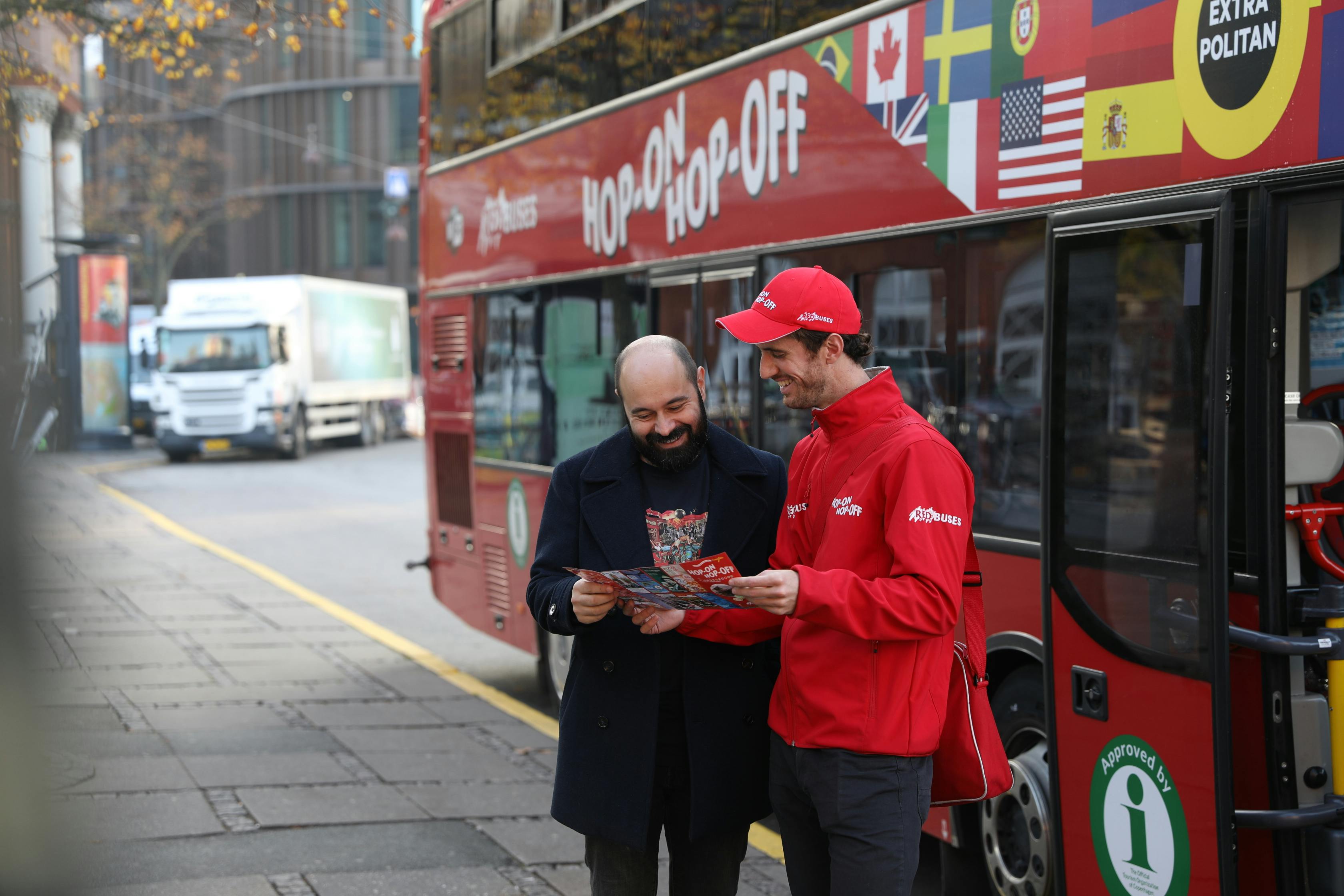 A tour guide and a tourist review a map beside a red hop-on hop-off bus with international flags on it.