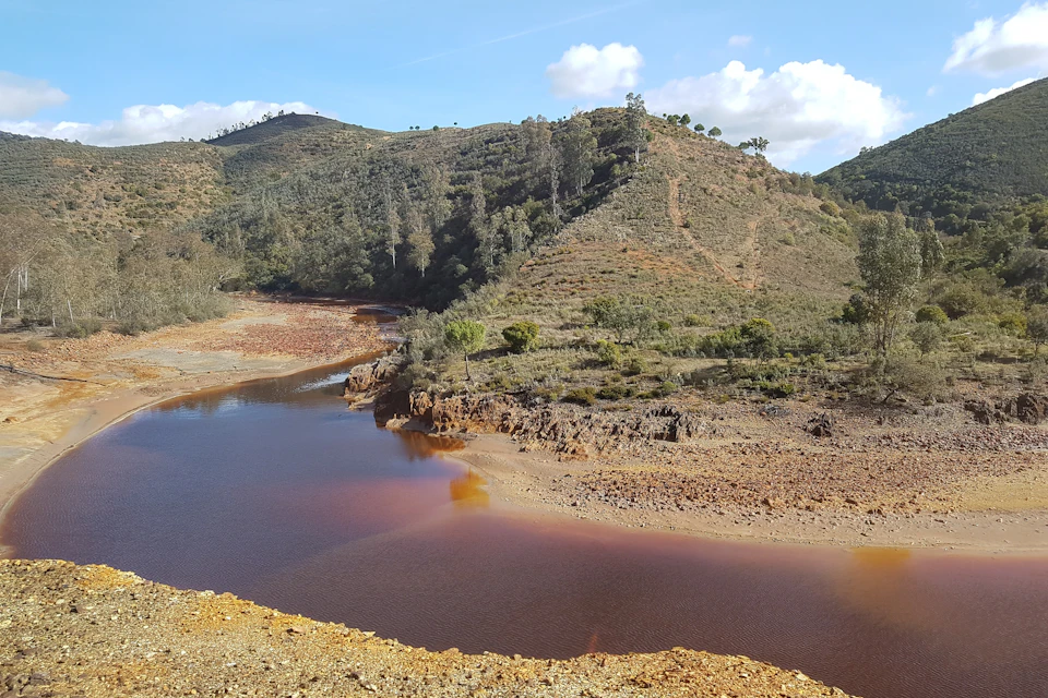Hillside with sparse vegetation surrounds a river with reddish-brown water reflecting the sky's light blue and cloud-speckled appearance.