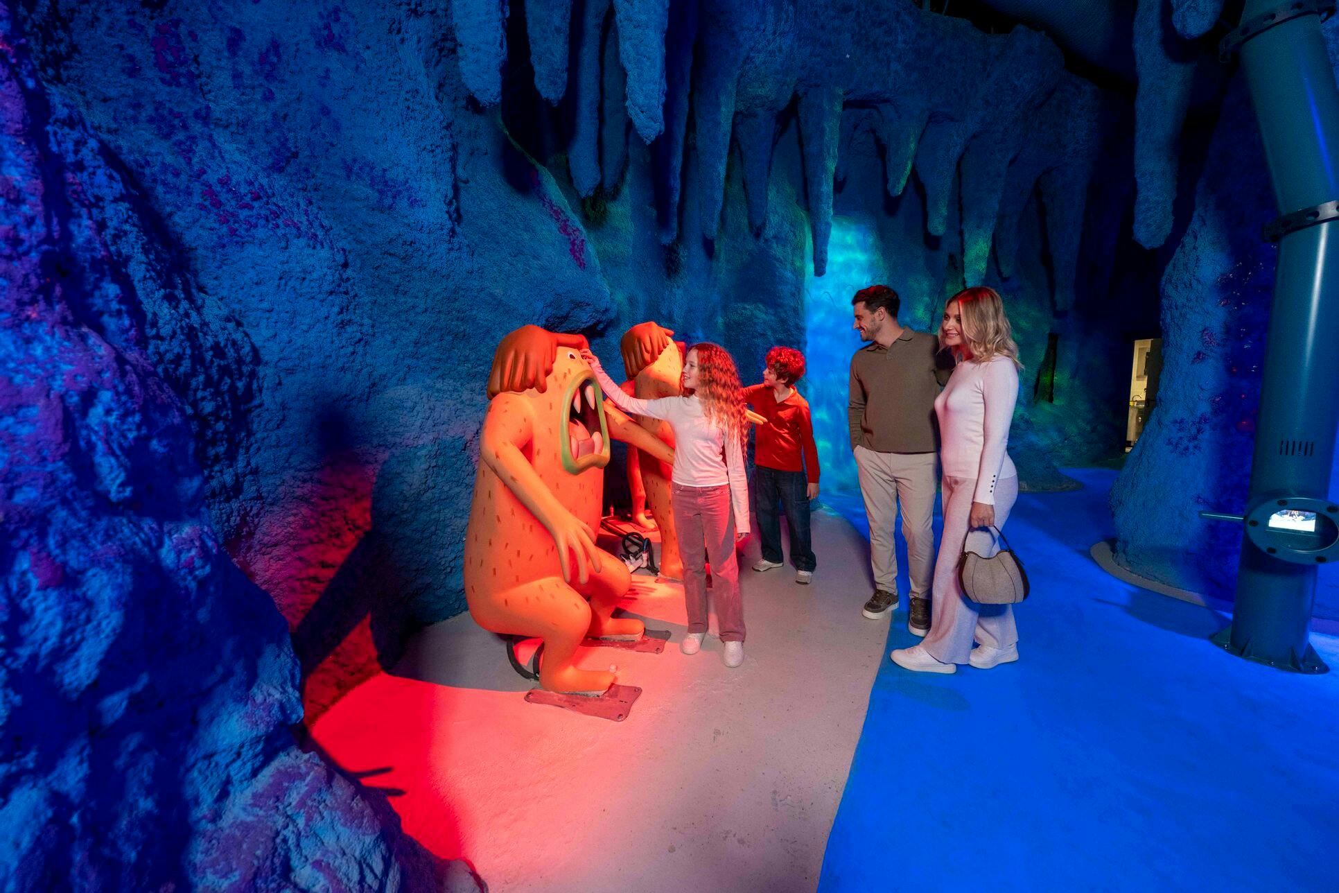 Mensen in interactie met kleurrijke beelden in een themakamer die lijkt op een grot met levendige blauwe verlichting en stalactieten.
