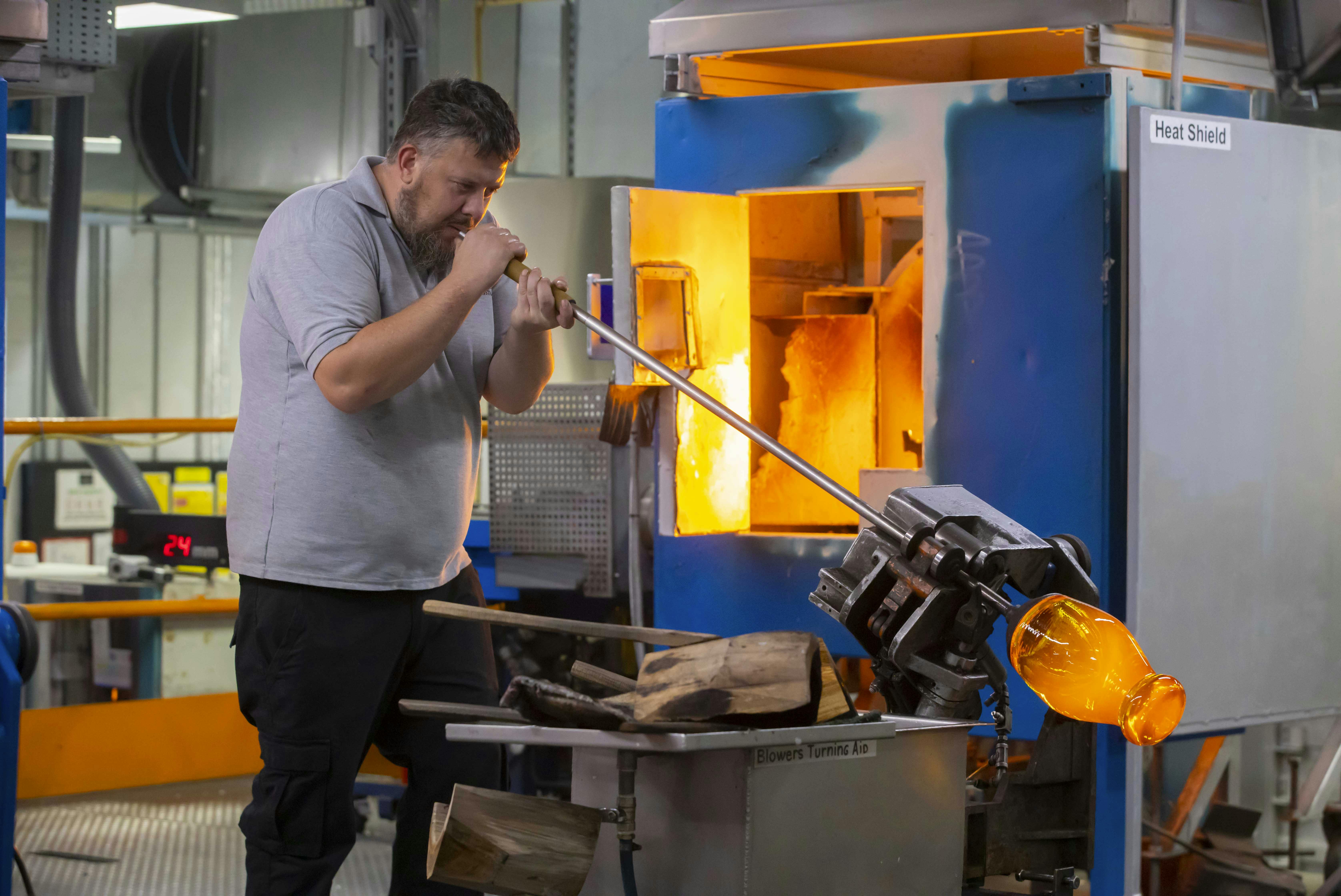 一名男子正在车间里用长管子吹玻璃，站在附近有工具和设备的玻璃炉旁边。
