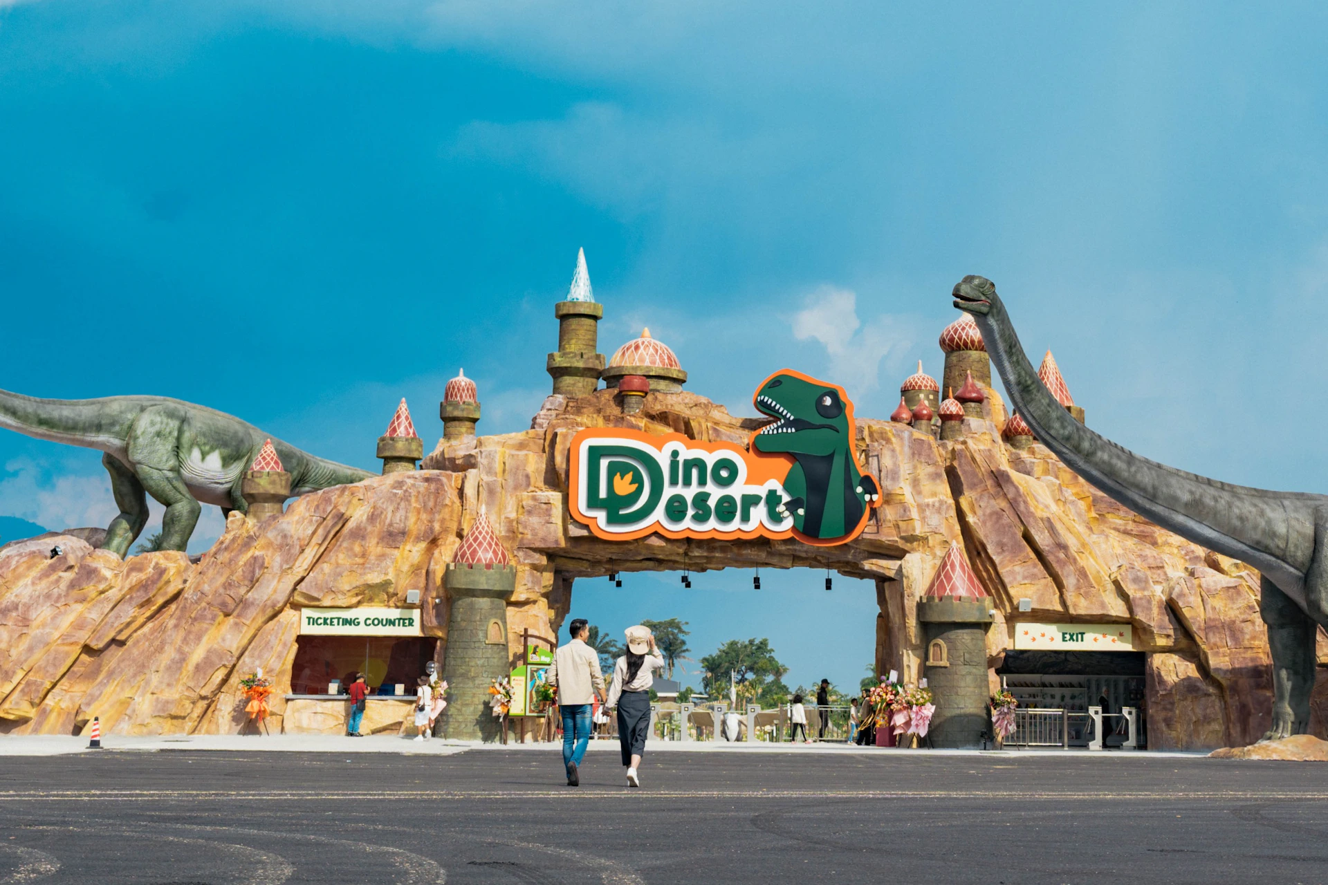 Entrance to Dino Desert amusement park, featuring dinosaur statues and a colorful sign under a blue sky.