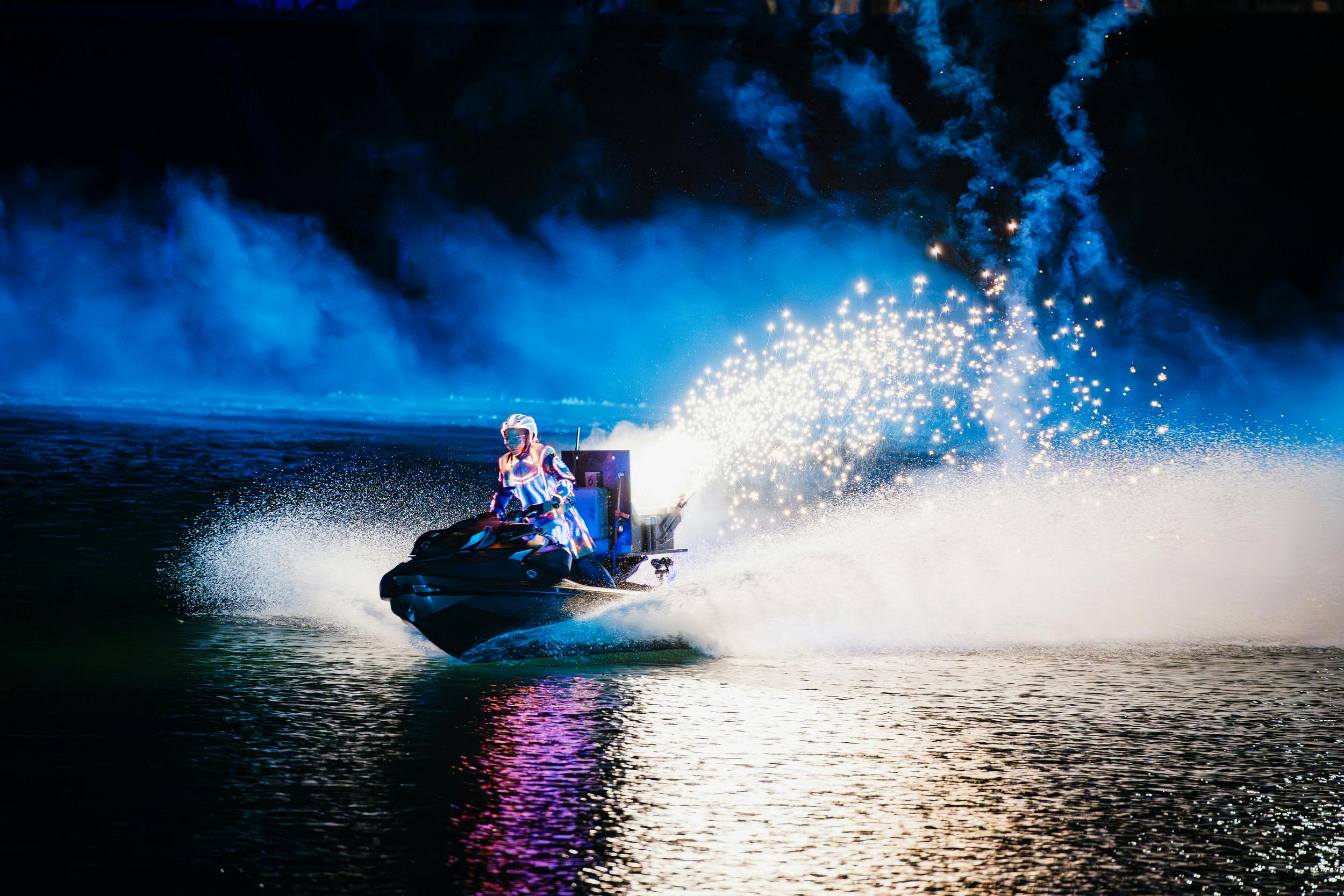 A person on a jet ski surrounded by bright sparks and smoke, creating a dynamic scene on water at night.