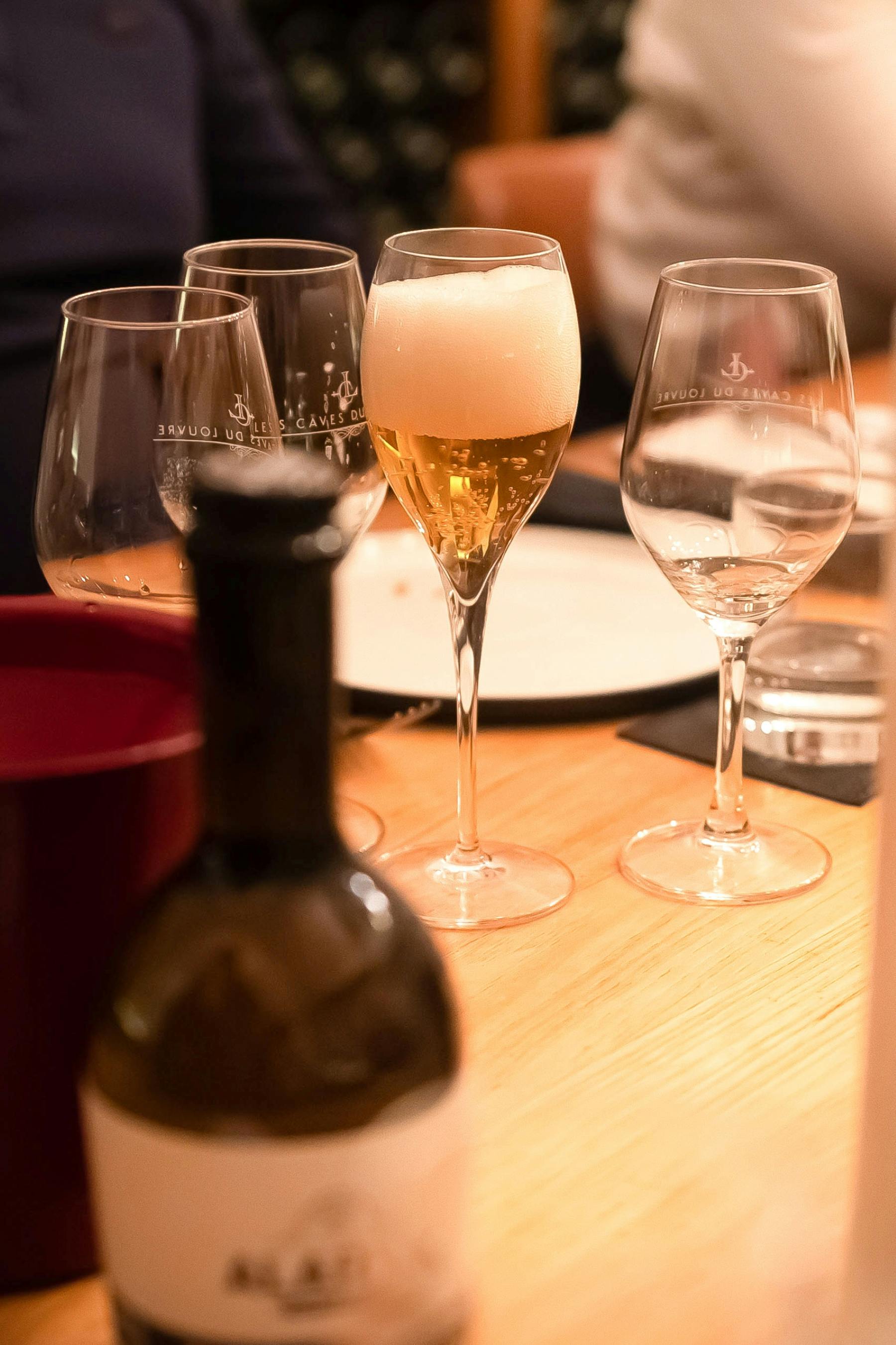 Обеденный стол с бокалами, в том числе с шампанским, и размытой бутылкой на переднем плане.