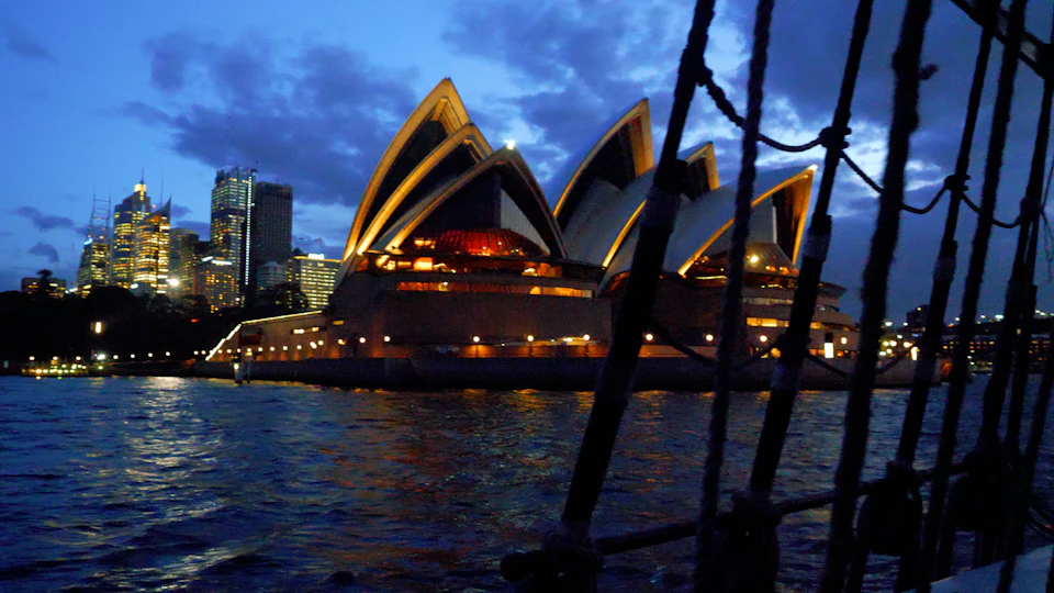 View of the Sydney Opera House at dusk, illuminated against the night sky, with water and a part of a boat in the foreground.