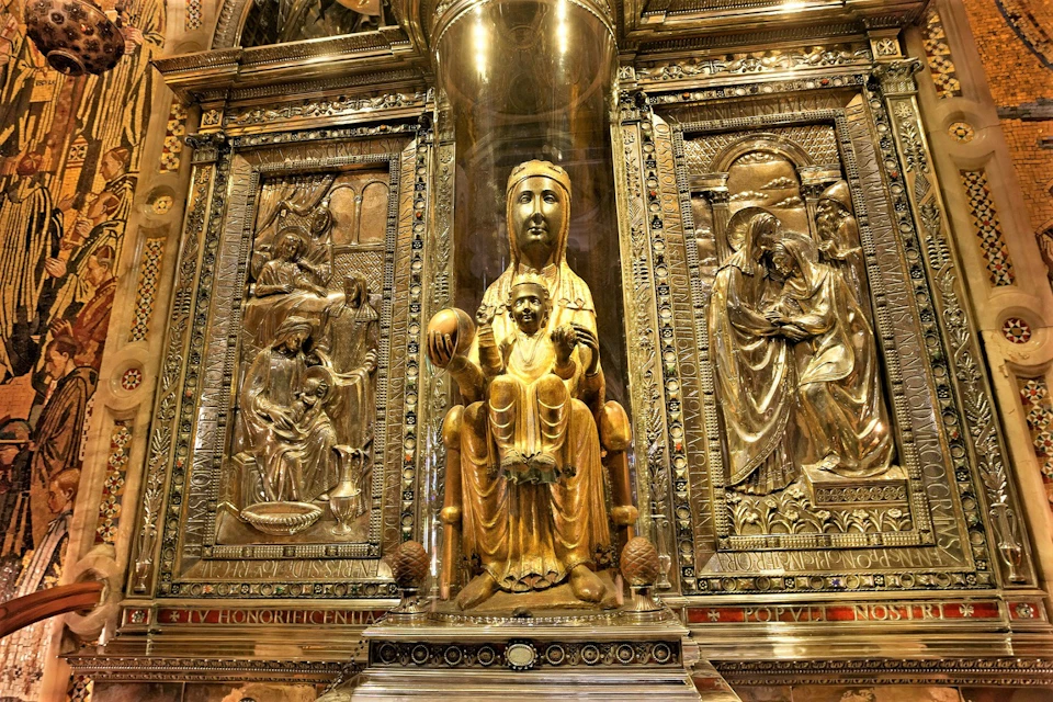 A golden statue of a seated woman with a child, flanked by intricate golden reliefs, within an ornately decorated area.