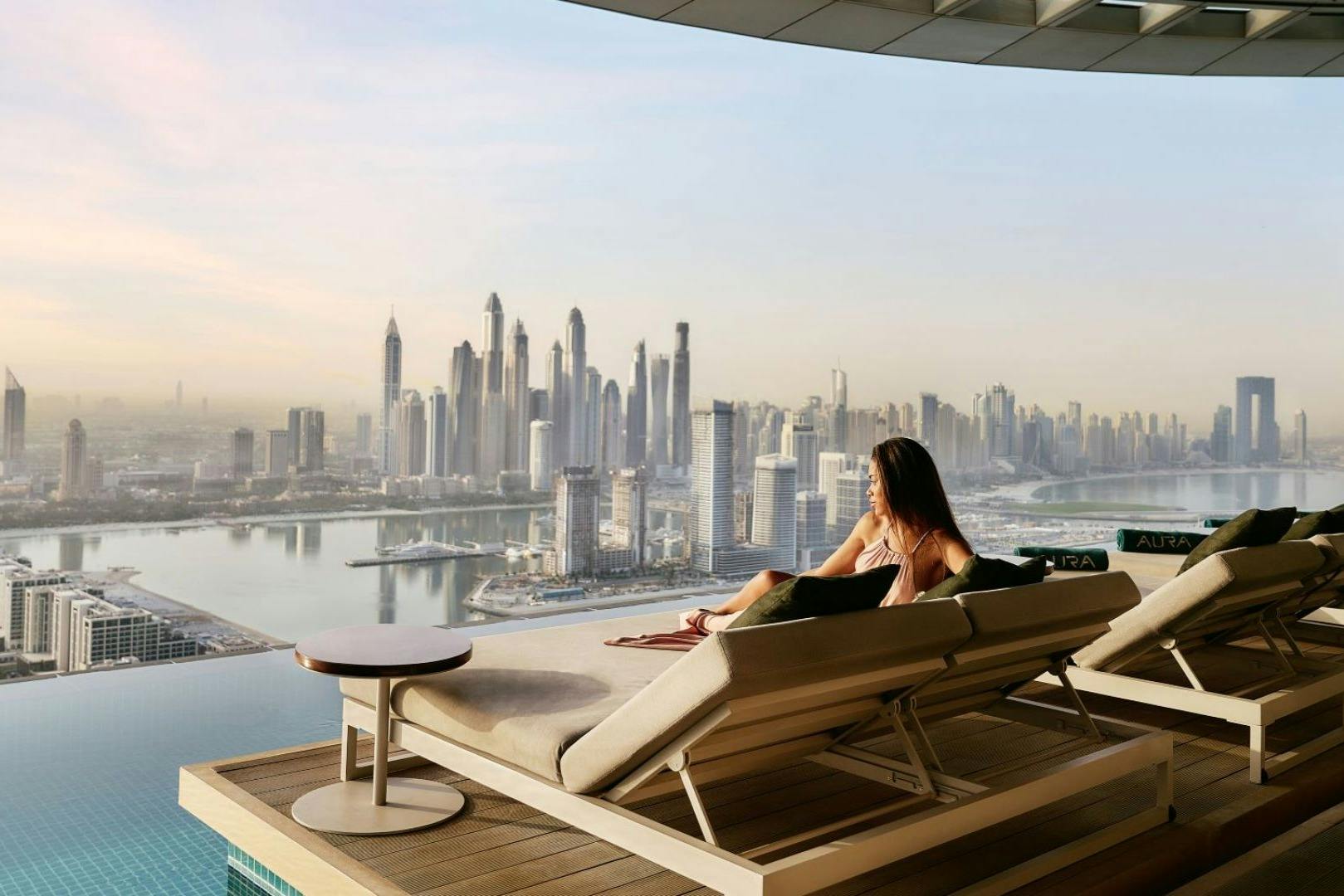 Žena odpočívá na lehátku u nekonečného bazénu s výhledem na panorama města s vysokými mrakodrapy a řekou.