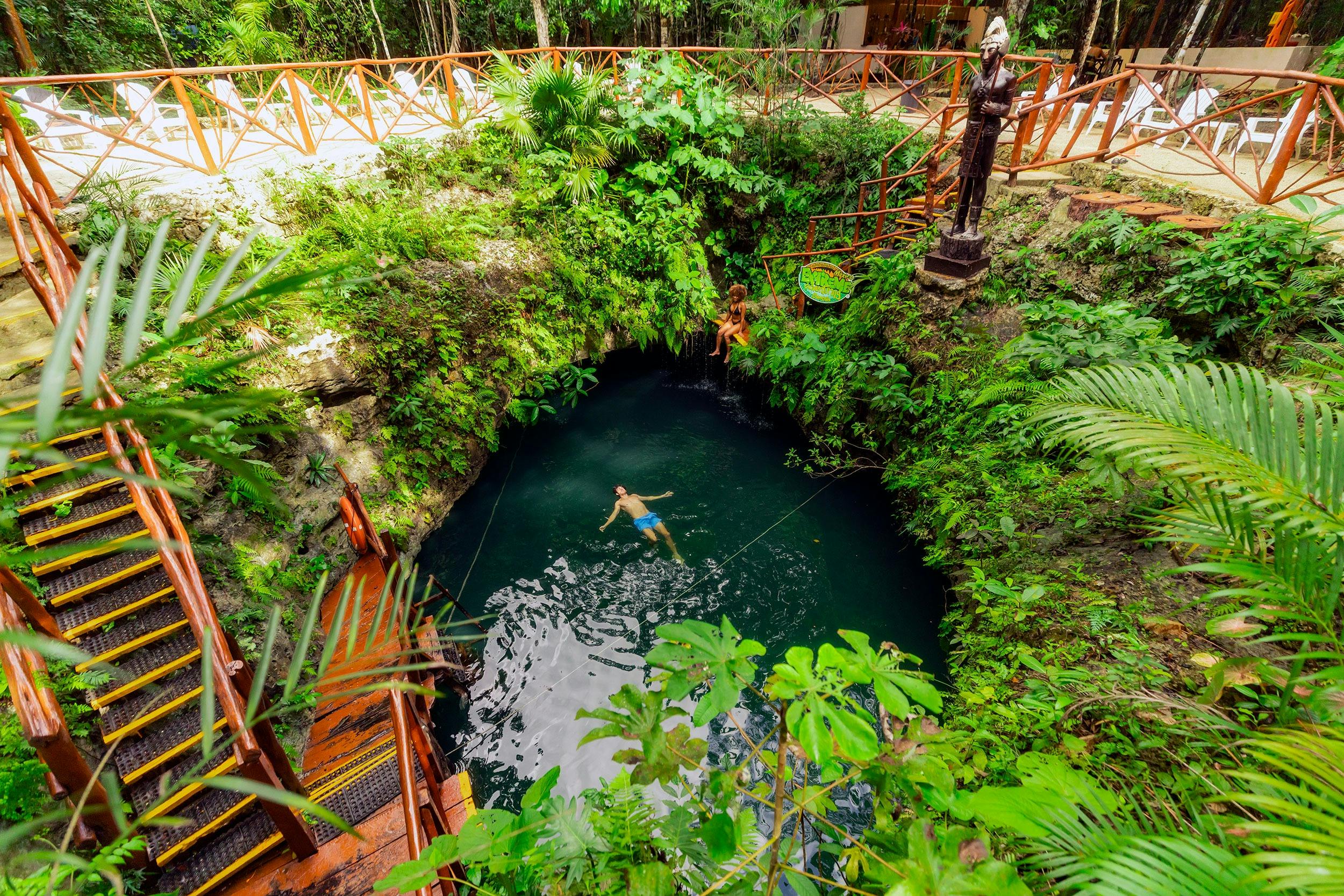 Una persona banya en un cenote natural envoltat d'una frondosa vegetació, baranes de fusta i estàtues en un entorn tropical.