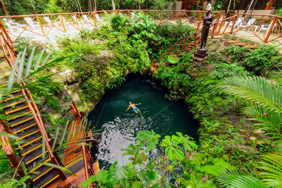 A person swims in a natural cenote surrounded by lush greenery, wooden railings, and statues in a tropical setting.