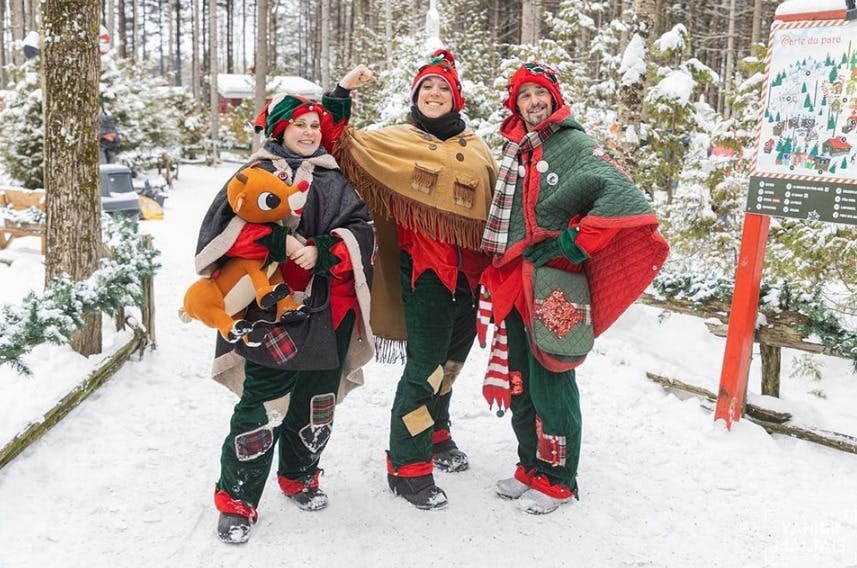 Drie mensen in feestkostuums staan in een besneeuwd bos, waarbij één een rendierspeeltje vasthoudt. Bomen en vakantiedecoraties op de achtergrond.