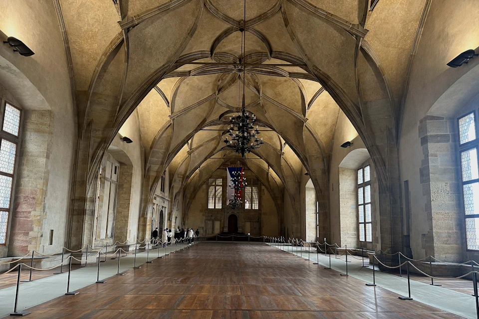 A spacious, historic hall with arched ceilings, wooden floors, chandeliers, large windows, and roped-off sections.