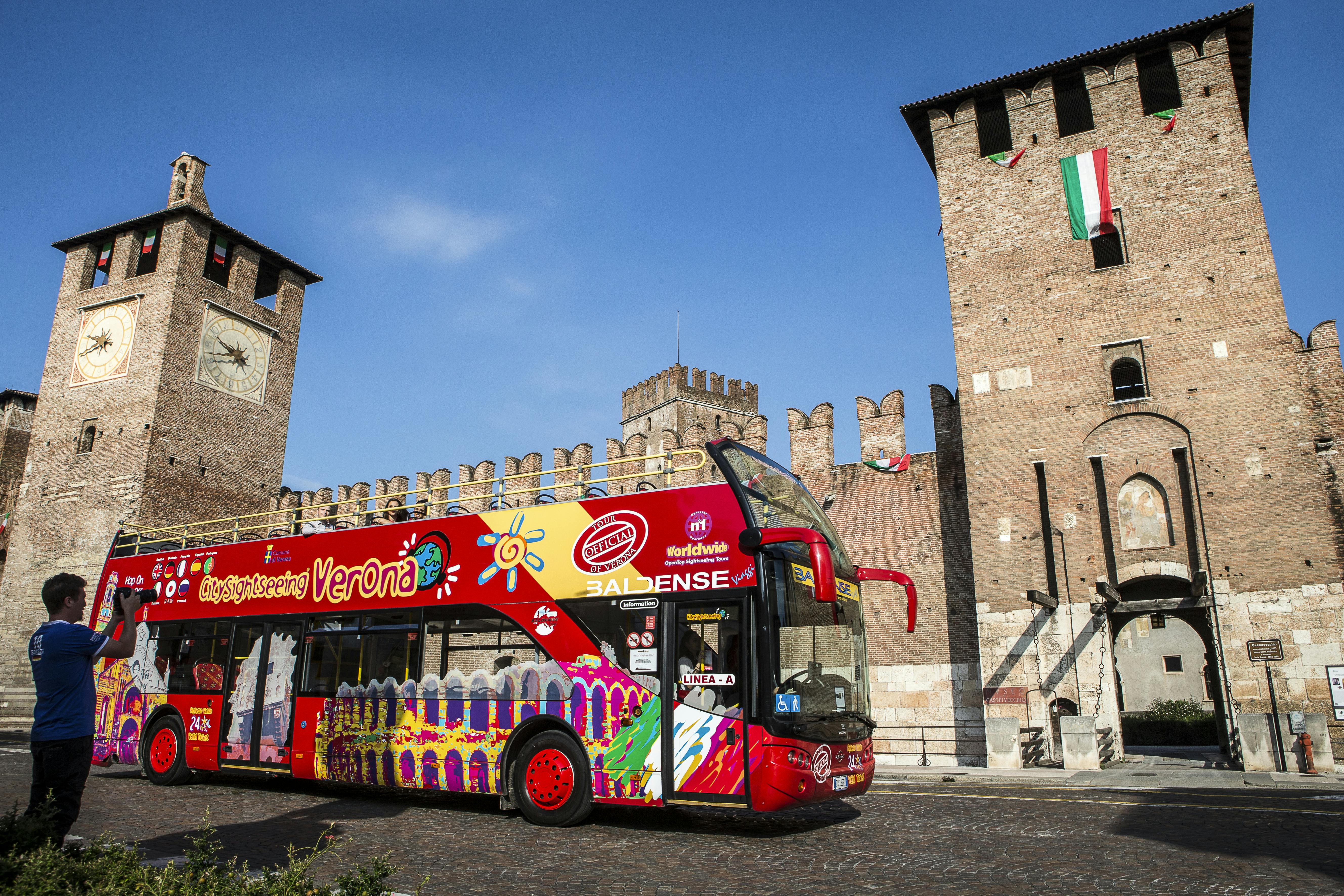 A red double-decker sightseeing bus in front of a historic brick building with towers and flags under a clear blue sky.
