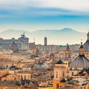 Skyline of a historic city with domed buildings, spires, and distant mountains under a blue, cloudy sky.